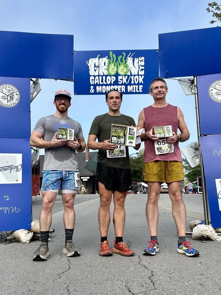 Three men standing under a blue arch at a race event, holding awards, with a sign that reads 'Girl Gallop 5K/10K & Monster Mile'.