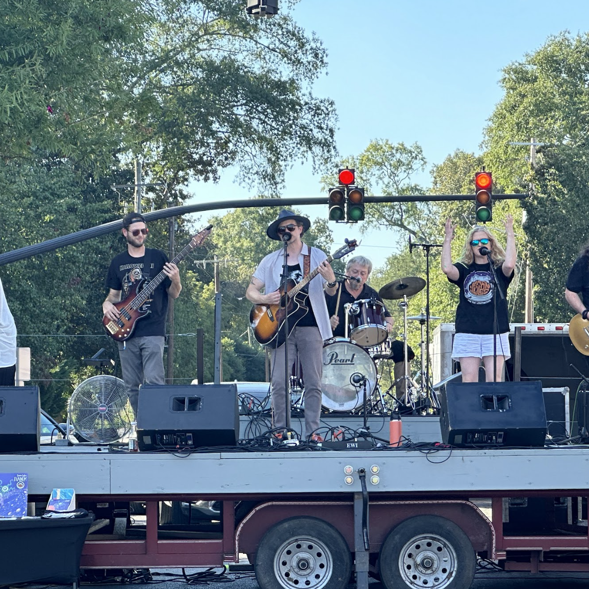A band performing live on an outdoor stage at the Green Eyes Festival with trees and a traffic light behind them. The band includes a guitarist singing into a microphone, a bassist, a drummer, and a female singer with sunglasses.