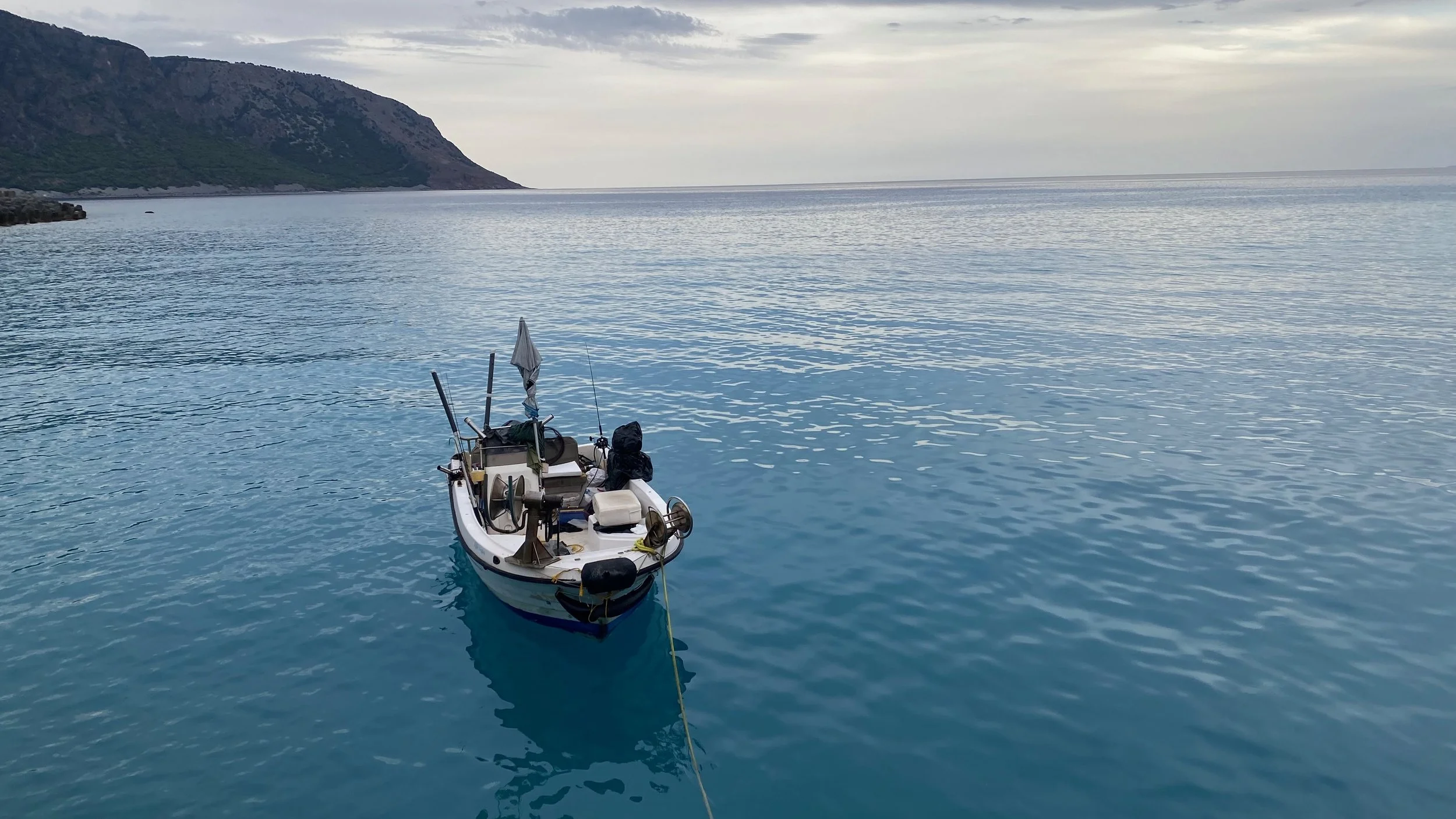 A small boat floating on calm blue water near a rocky shoreline and mountainous coast in the distance, under a cloudy sky.