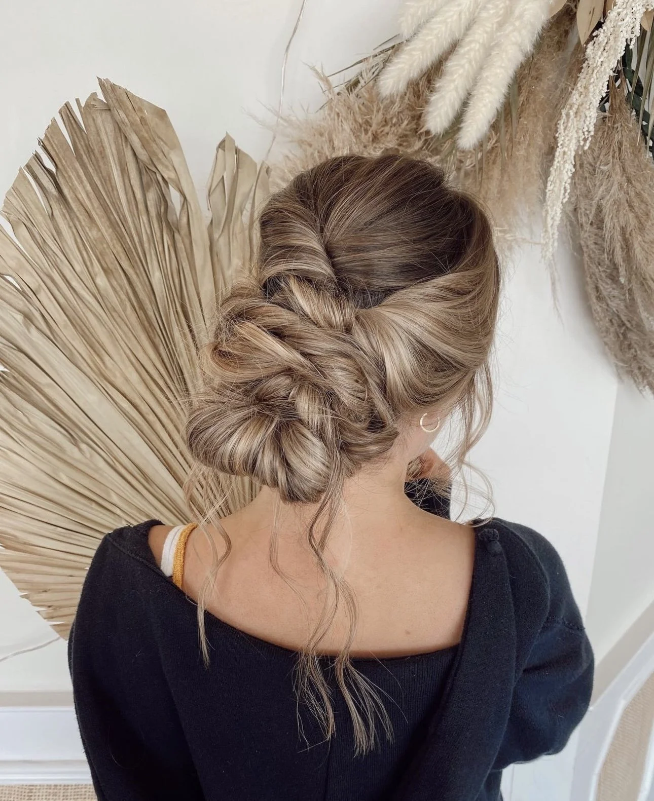 Back view of a woman with blonde hair styled in an elegant twisted updo, wearing a black top, in a room with decorative dried plants and pampas grass.