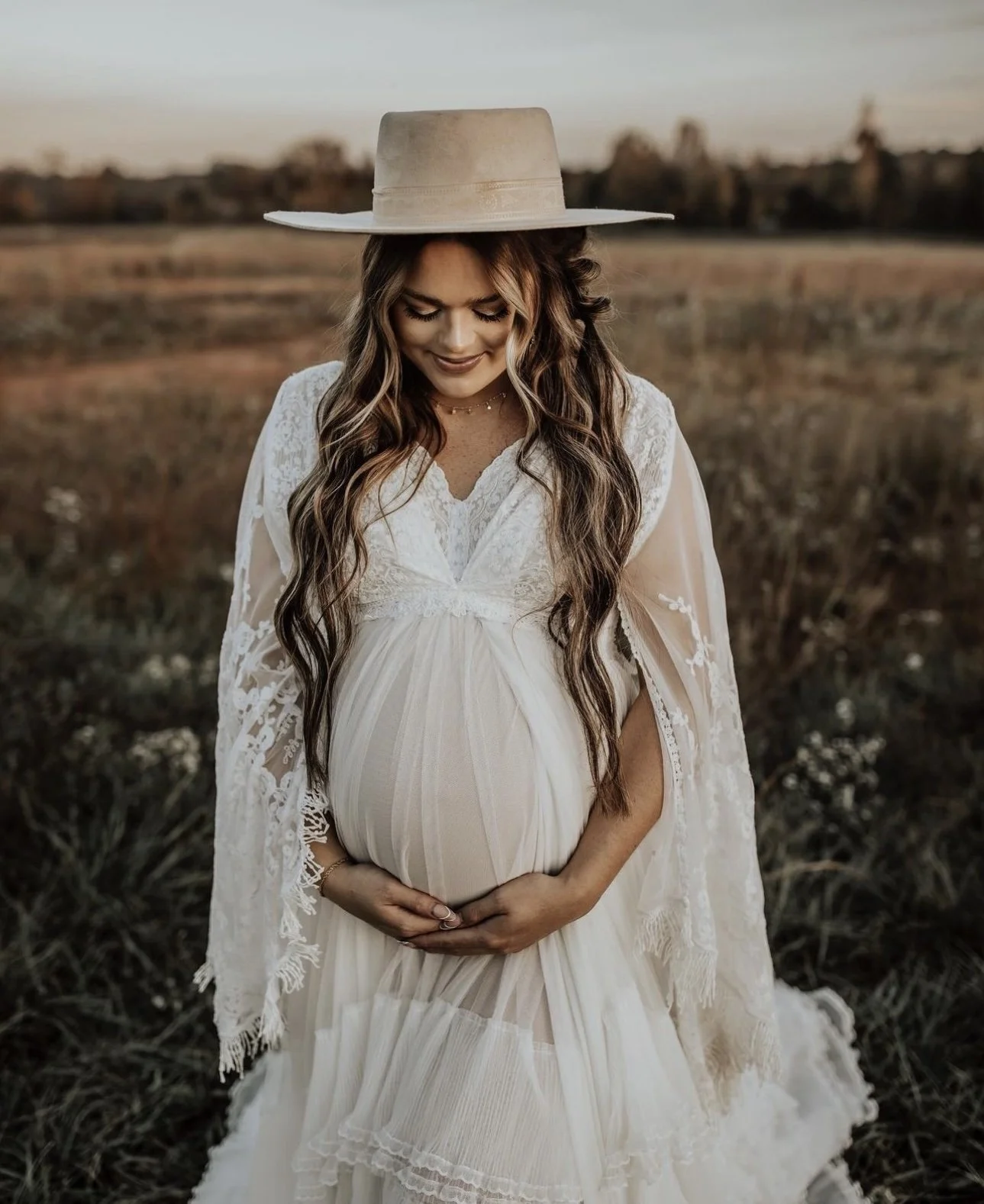 A pregnant woman in a white lace dress kneeling in a field during sunset, wearing a wide-brimmed hat and smiling gently while holding her baby bump.
