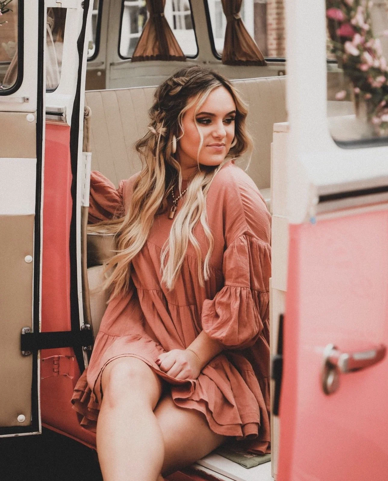 A young woman with long, wavy blonde hair sitting inside a vintage camper, wearing a pink bohemian dress with puffed sleeves, looking to her left with a relaxed expression.