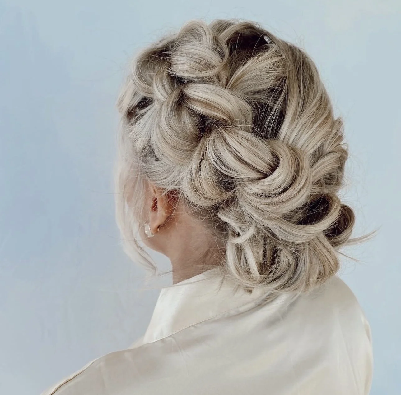 Elegant blonde hair styled in an intricate updo with twists and curls, worn by a woman in a cream-colored outfit against a light blue background.