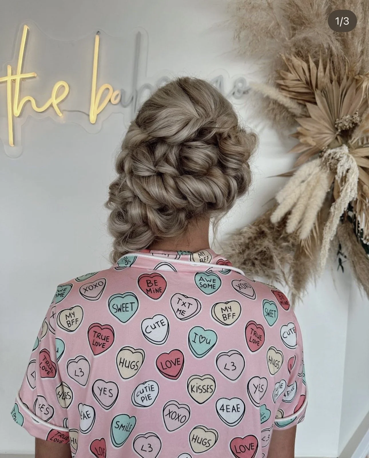 Back view of a woman with curly, braided silver hair wearing a pink shirt with colorful heart patterns and messages like "be mine," "sweet," and "love". Behind her, a wall features a neon sign and a dried floral arrangement.