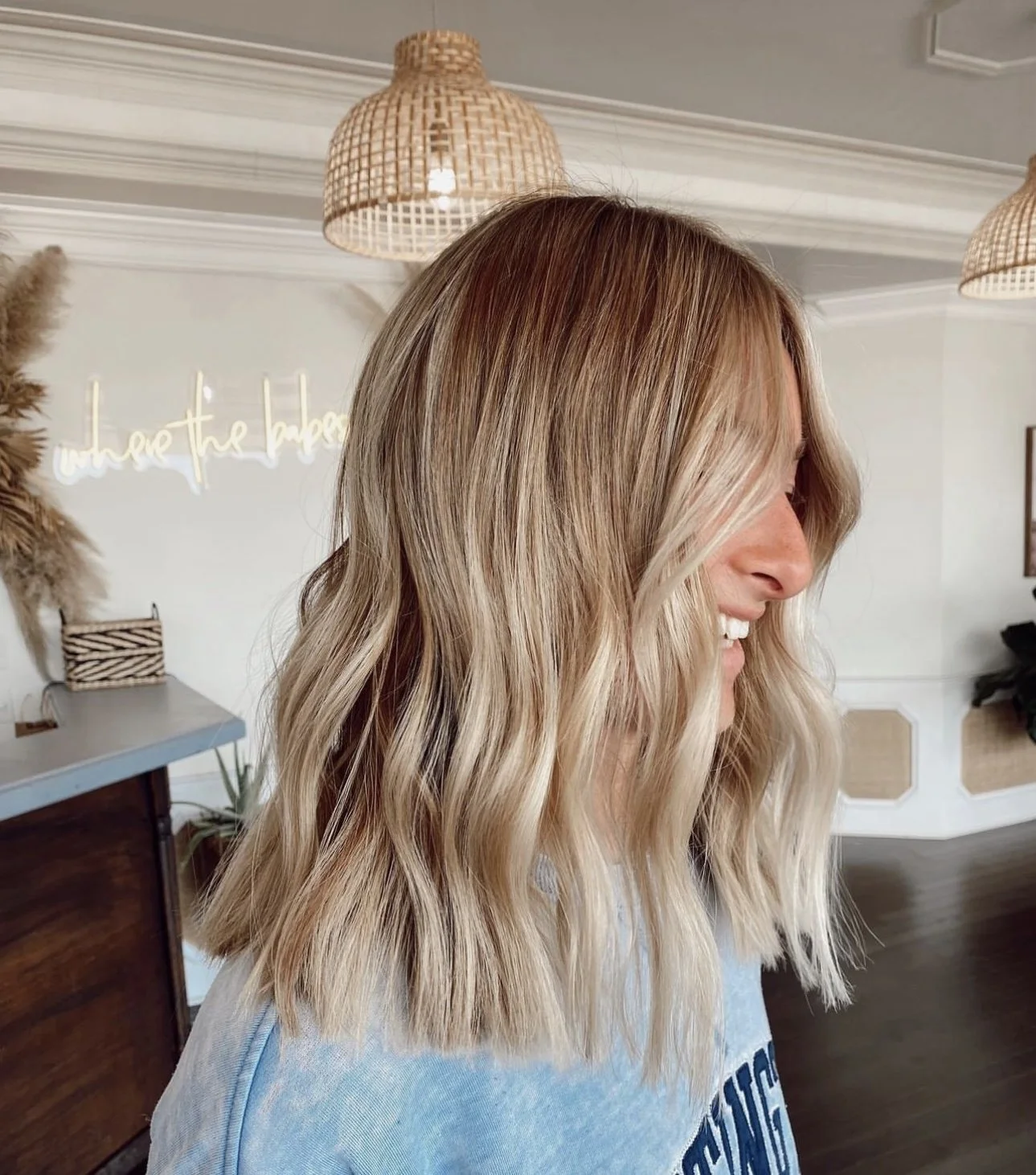 A woman with blonde, wavy hair smiling in a modern, well-lit room with woven pendant lights and decorative plants.