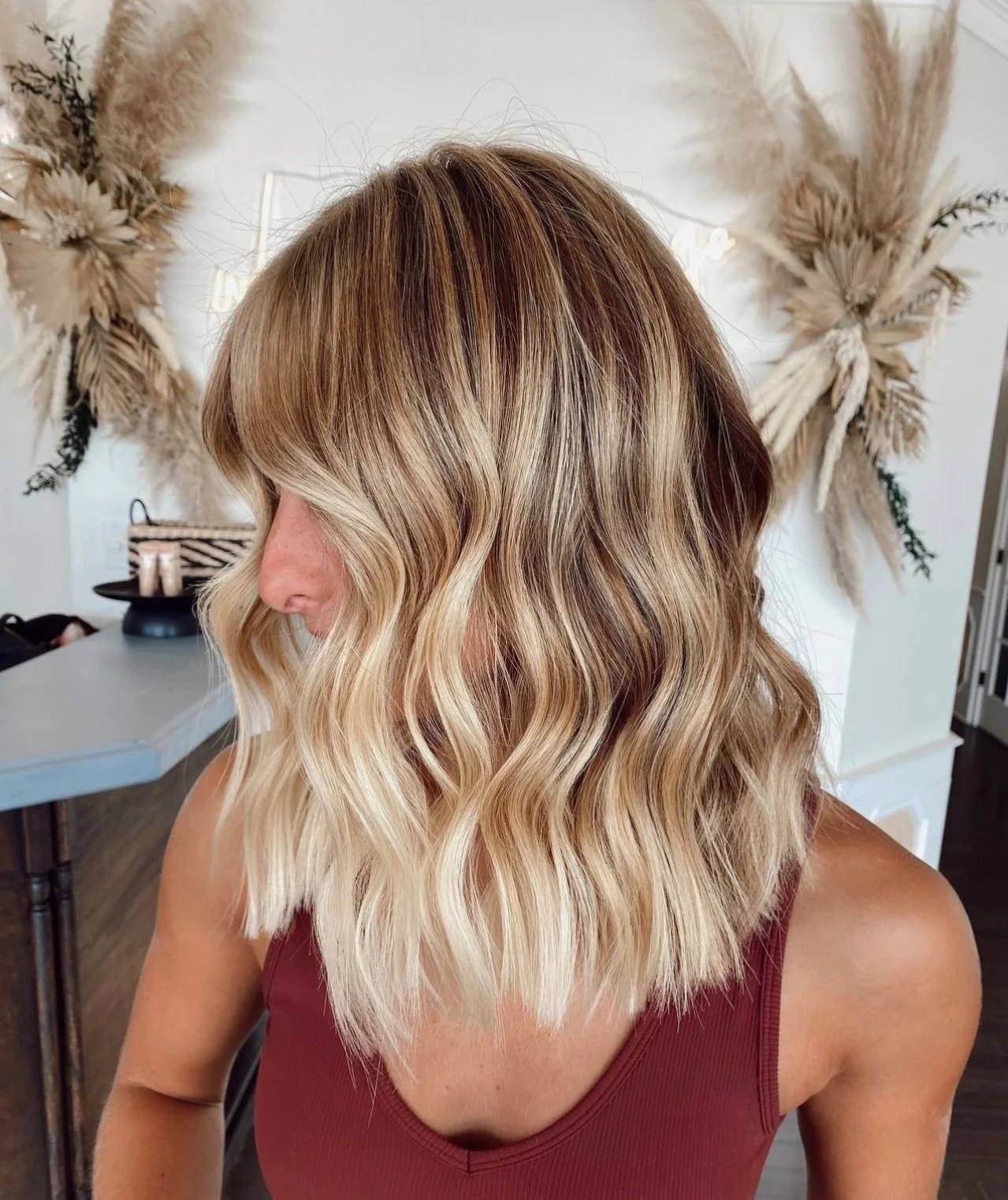 Woman with shoulder-length wavy blonde hair, wearing a maroon tank top, in a room with dried floral wall decor in the background.