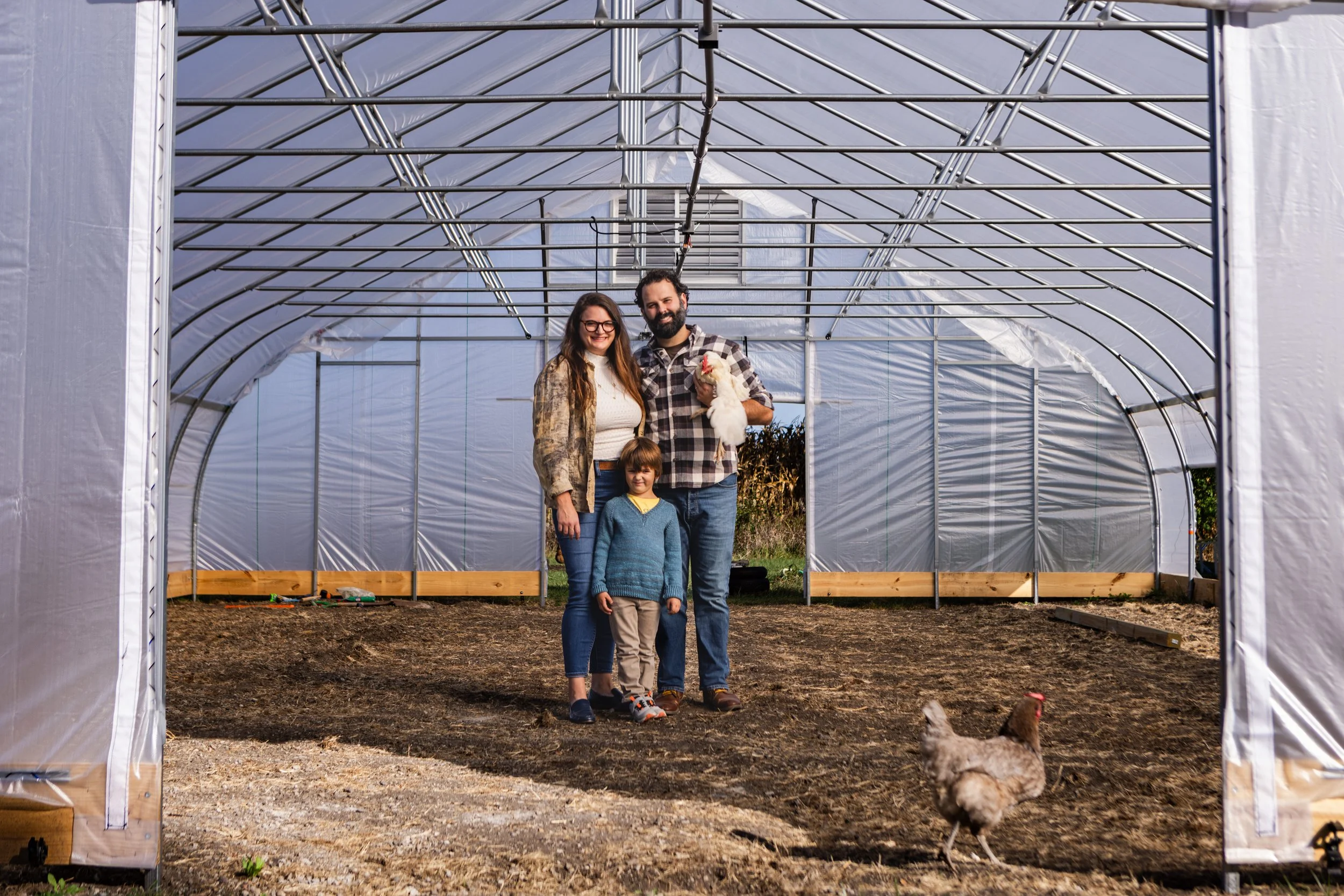 Family standing inside a greenhouse with a chicken and a rooster outside.