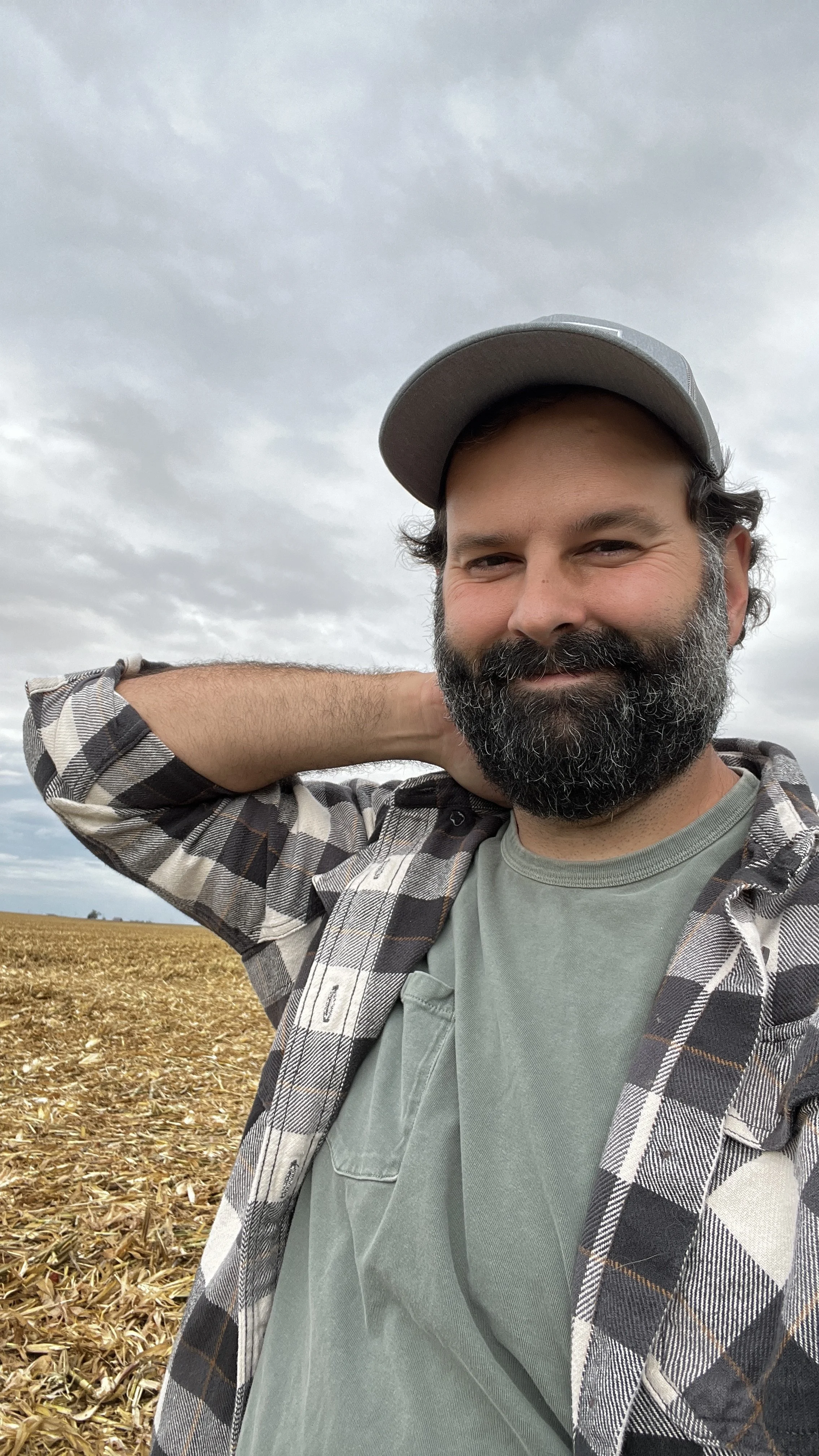 A man with a beard and gray cap smiling outdoors in a field of harvested corn with cloudy sky in the background.