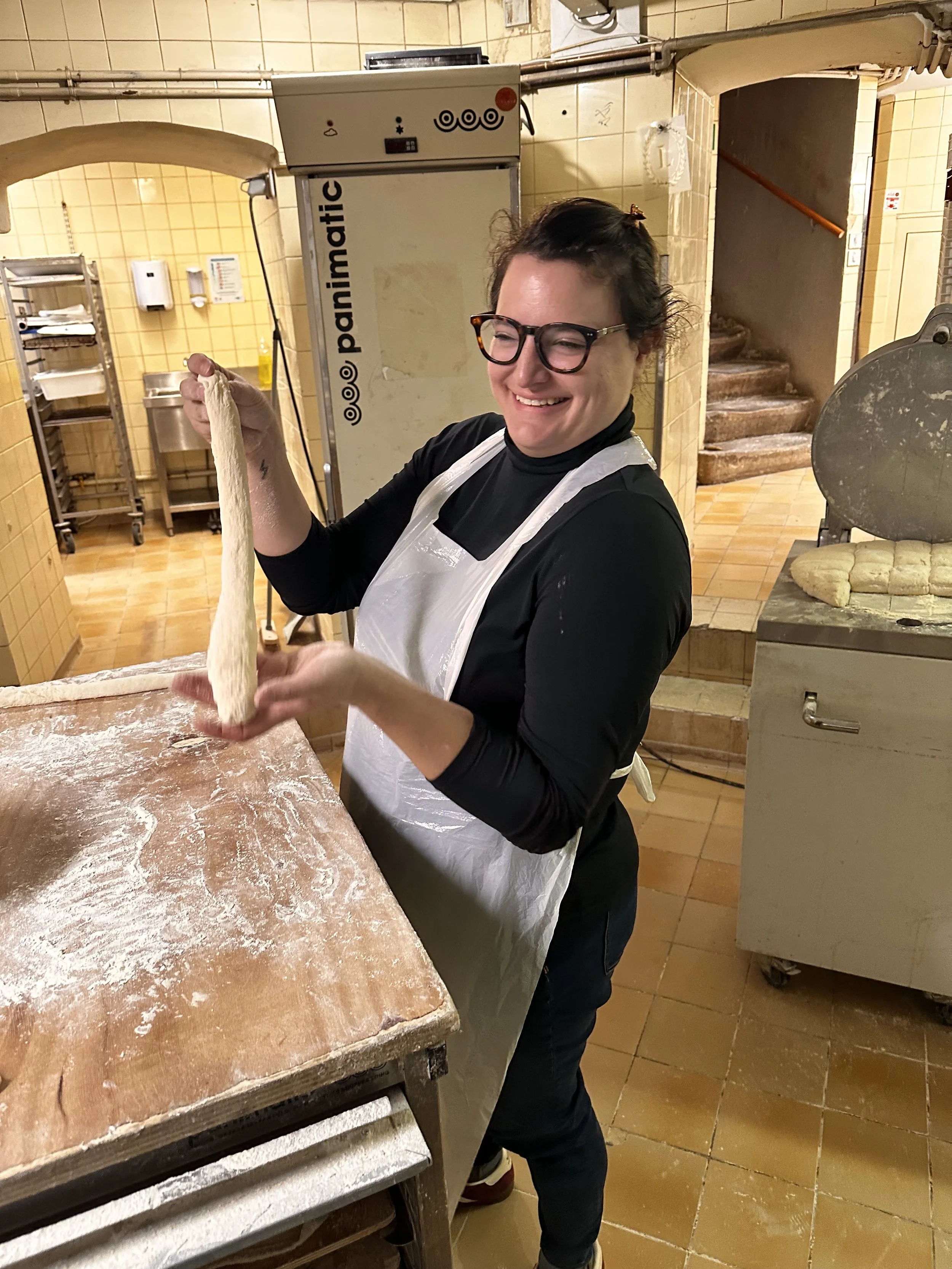Woman with glasses and an apron holding a long piece of dough in a bakery kitchen.