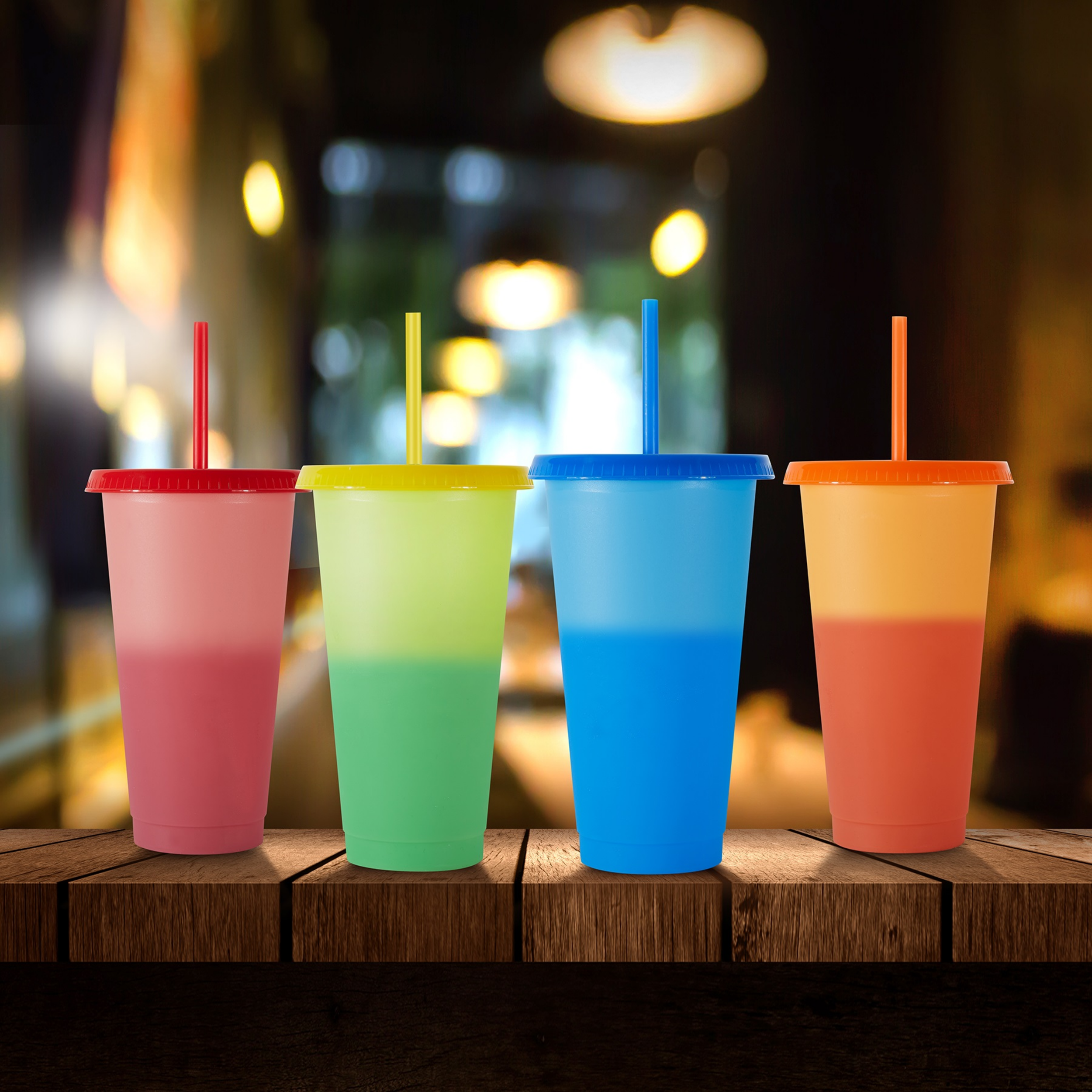 Colorful cups with straws on a wooden table, blurred background with warm lights.
