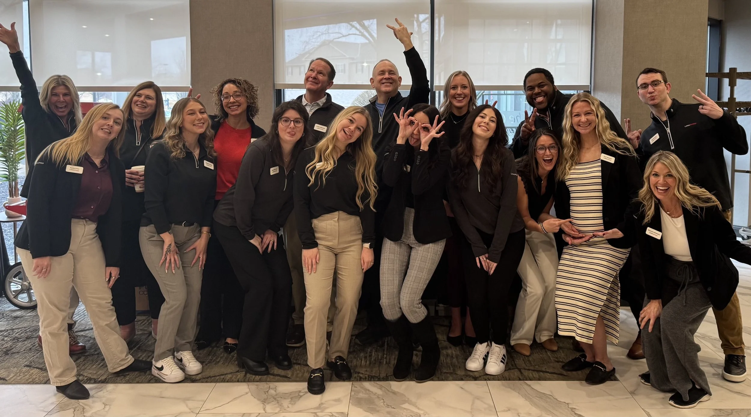 A group of people, posing indoors with smiles and making playful gestures. They are dressed in business casual attire and appear to be at a professional or social event.