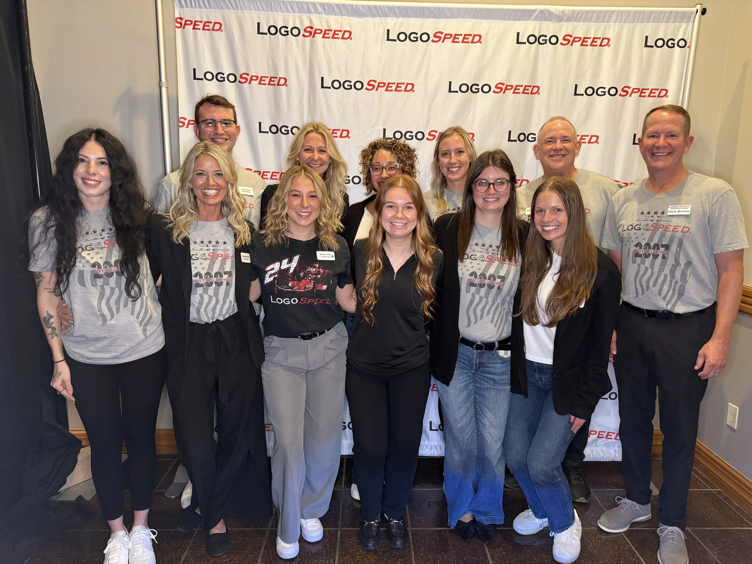 Group of eleven people posing in front of a backdrop with 'LOGO SPEED' written repeatedly, some wearing matching gray t-shirts with racing graphics, others in black or gray casual clothes, indoors on a wooden floor.