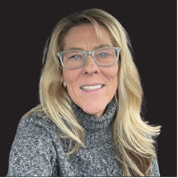 A woman with long blonde hair wearing glasses and a gray sweater, smiling at the camera against a black background.