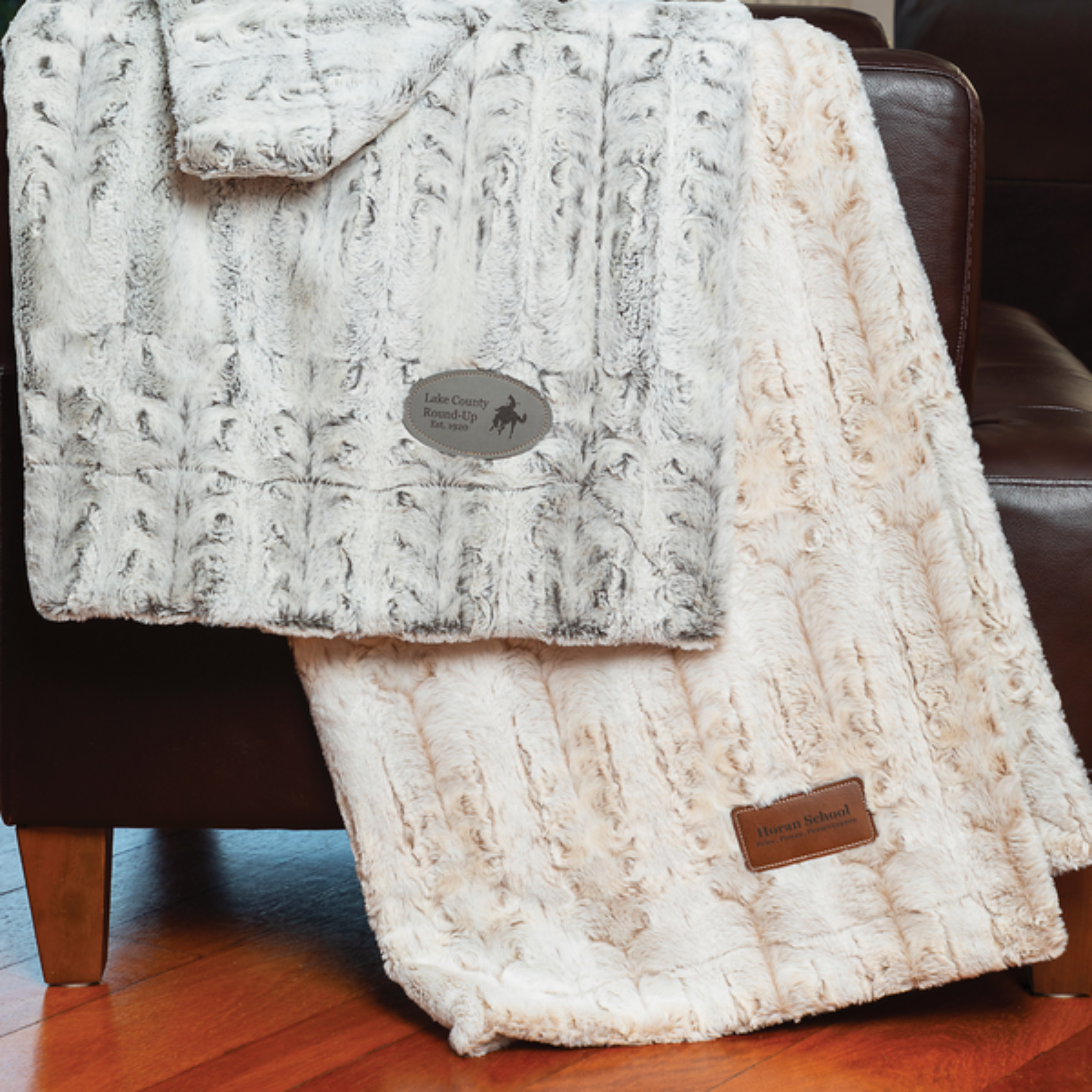 Two plush throw blankets draped over a dark leather armchair. One is light gray with a pattern, the other is cream-colored with a leather label.