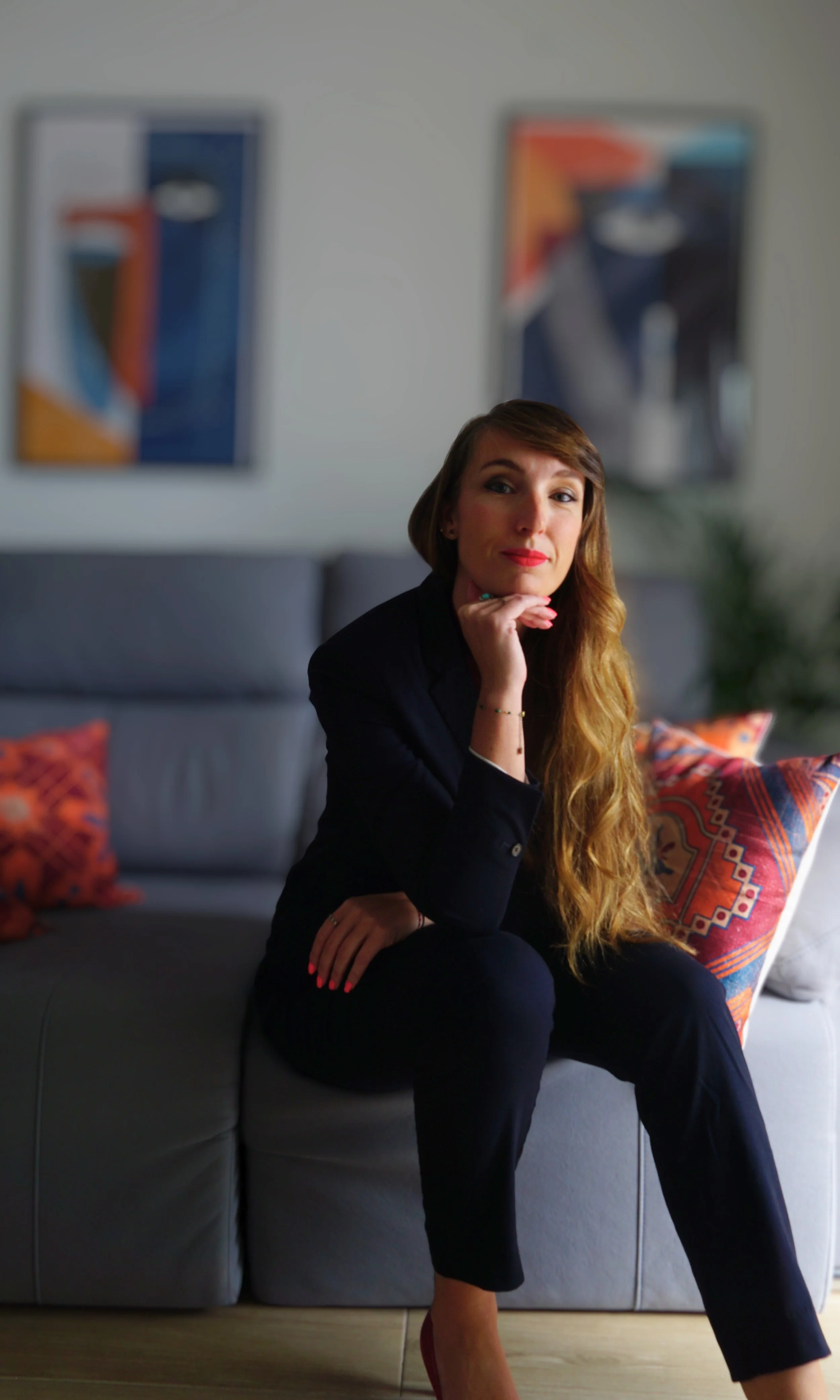 Picture of Valerie Leroux: A woman with long red hair, wearing a black suit, sitting on a gray couch with colorful pillows, resting her chin on her hand, in a living room with abstract artwork on the wall.