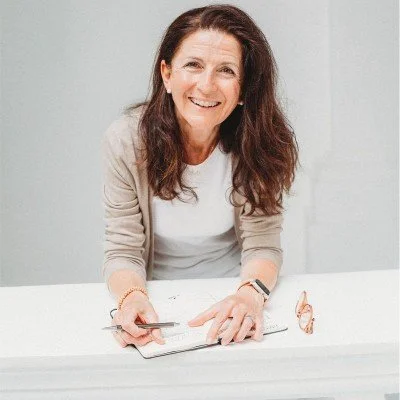 A smiling woman with long brown hair sitting at a white table, holding a pen and a notebook, with a pair of glasses beside her.
