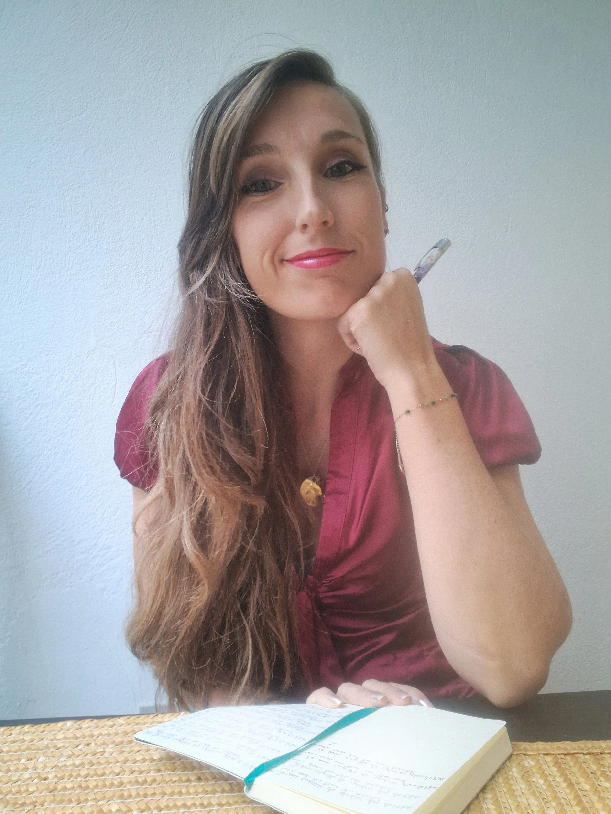 Photo of Valerie Leroux, a woman with long brown hair, wearing a maroon blouse, sitting at a table with a notebook, holding a pen near her face and looking at the camera with a slight smile.