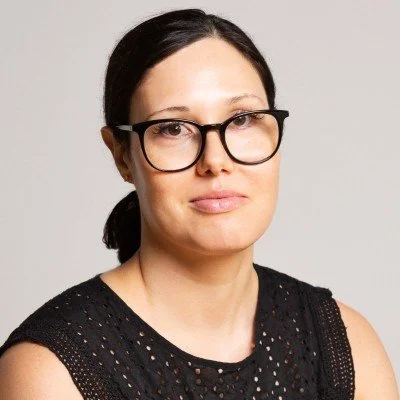 Portrait of a woman with dark hair, wearing glasses, and a black sleeveless top with a laser cut design, against a light gray background.