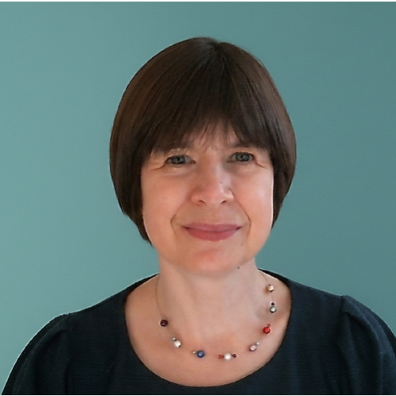 Sinead profile picture: A woman with short brown hair and a gentle smile, wearing a black top and a colorful beaded necklace, standing in front of a teal background.