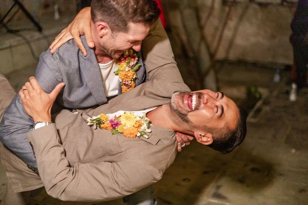Two grooms dancing and laughing together at their wedding reception, with one dipping the other.