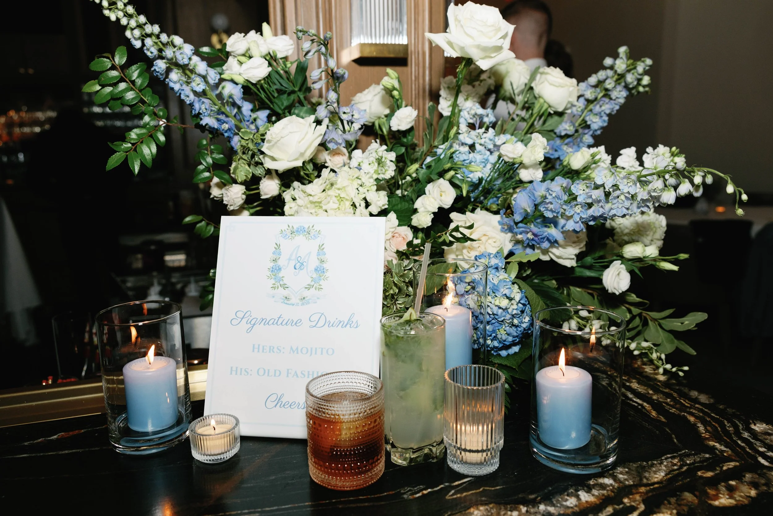 Blue and white wedding bar arrangement with fresh flowers, a His and Hers drink menu sign, and signature cocktails.
