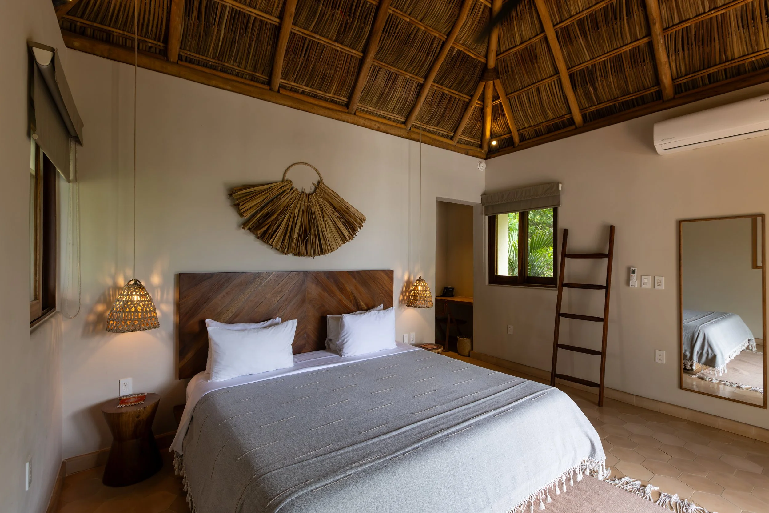Interior of a cozy room with a large sliding glass door leading to a balcony with a hammock, surrounded by trees and natural scenery outside. The room has a bed with a white blanket, a small stool, and decorative lighting creating a warm ambiance.