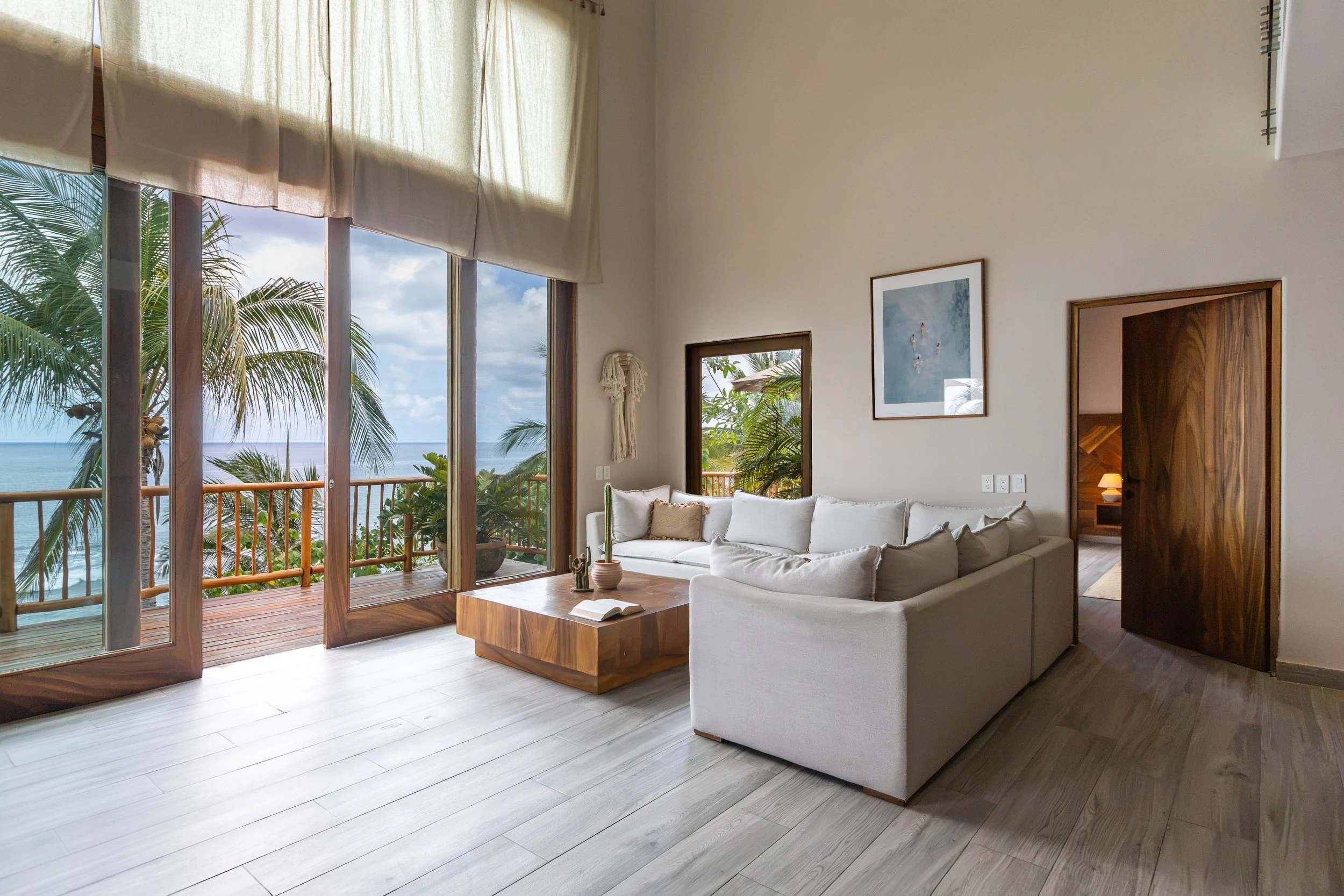 Living room with a white sofa, beige pillows, wooden coffee table, sliding glass door leading to a balcony with tropical plants, window with view of outdoors, framed artwork on beige wall.