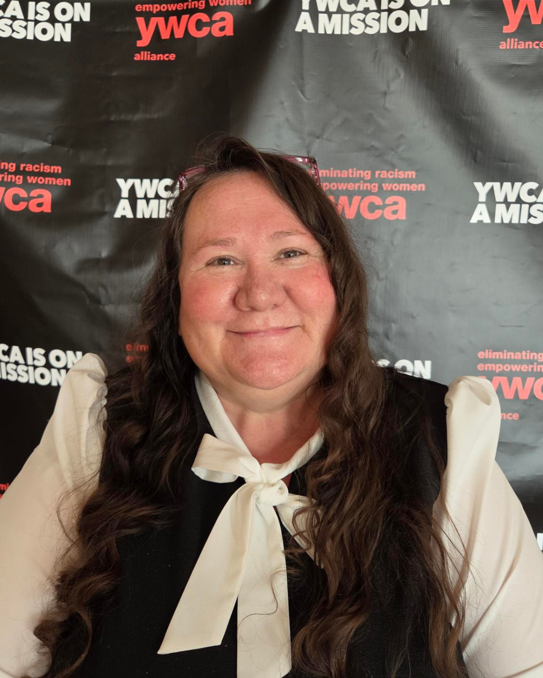A woman with long, wavy brown hair smiling in front of a black YWCA backdrop. She is wearing a cream-colored top with a bow tie at the collar and a black vest.