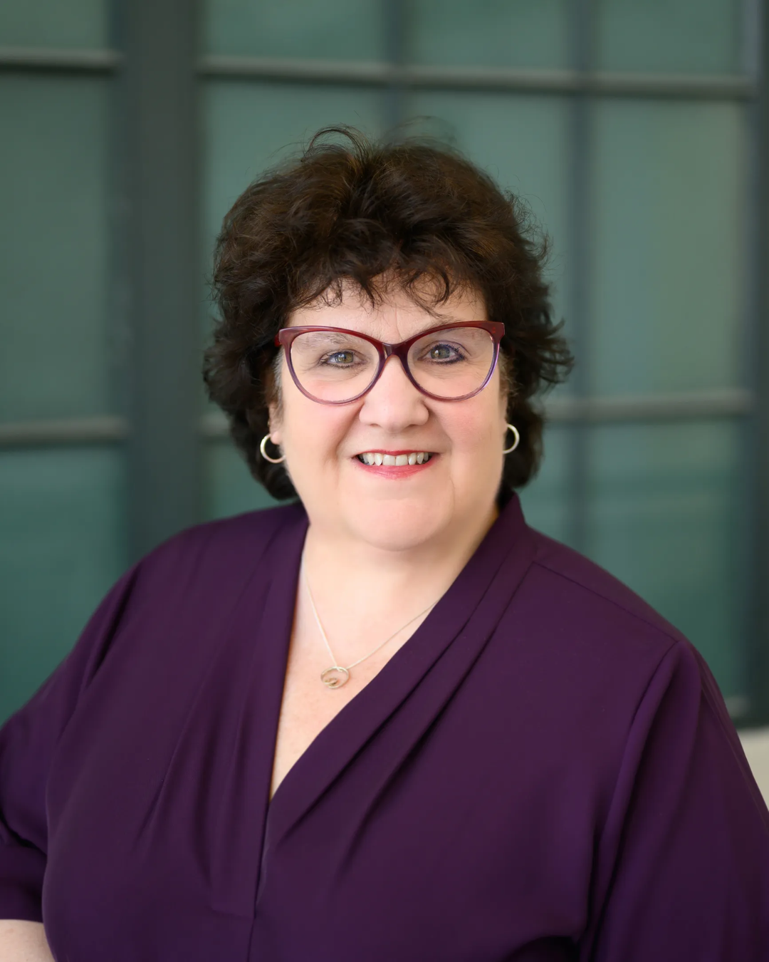 Portrait of a woman with curly dark hair, glasses, and a purple top, smiling in front of green window panels.