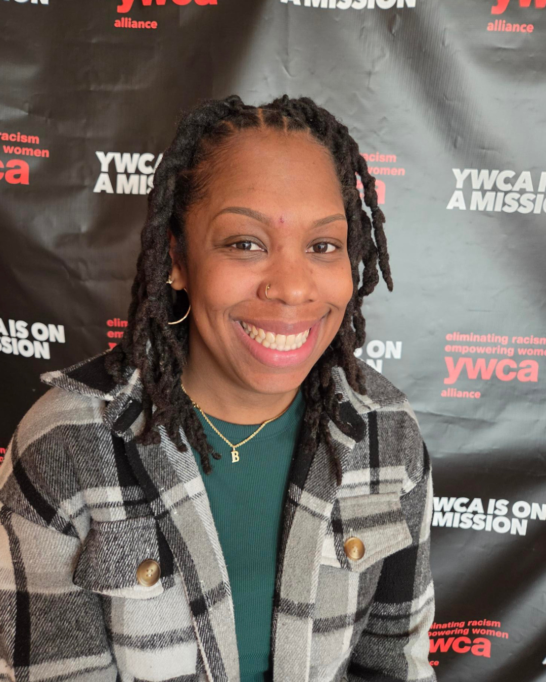 A smiling woman with dreadlocks and a nose ring, wearing a plaid jacket and a green top, standing in front of a black backdrop with red and white text that reads 'YWCA' and 'eliminating racism empowering women'.