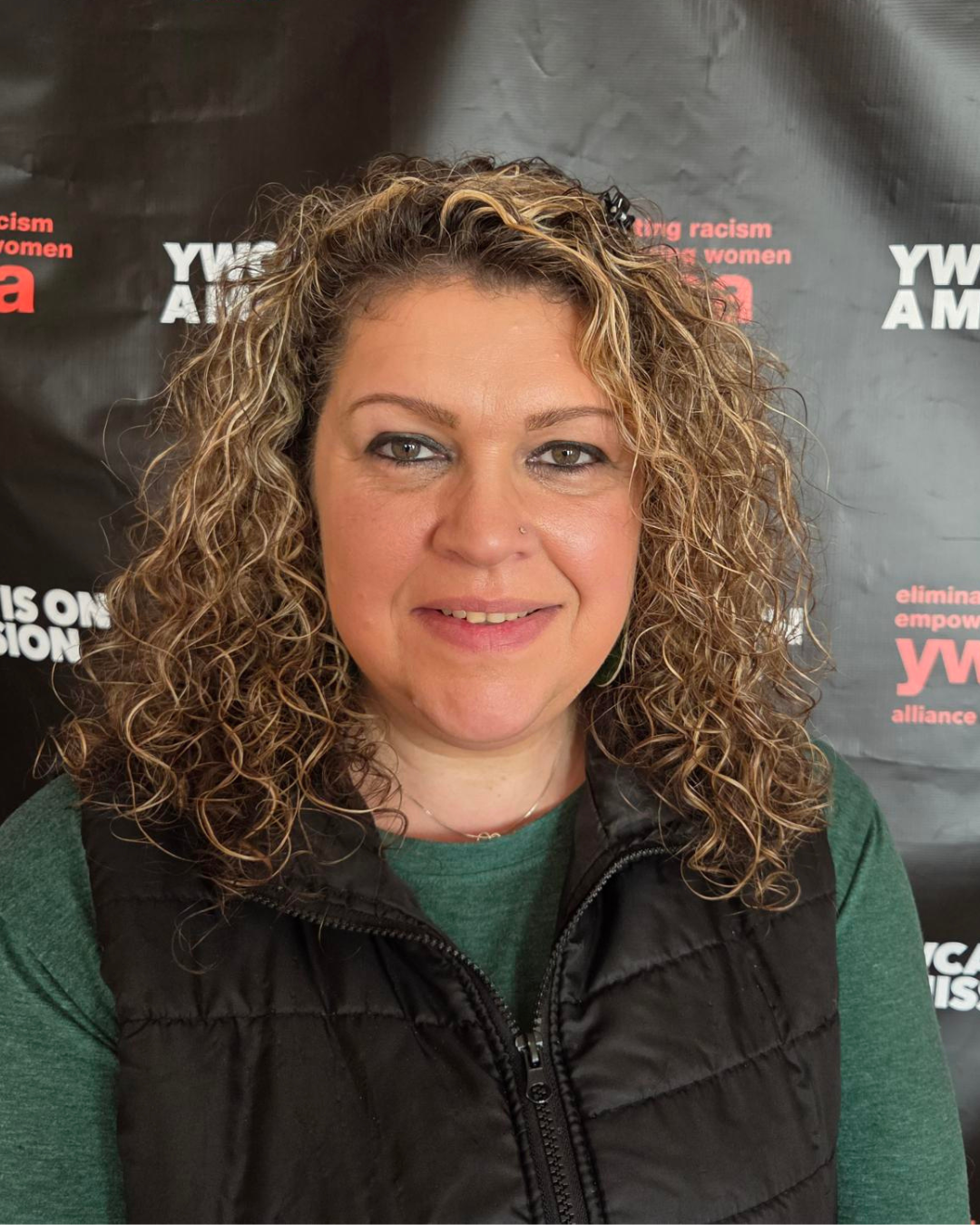 A woman with curly blonde hair smiling at the camera, wearing a dark vest over a green shirt, in front of a black background with white and red text.