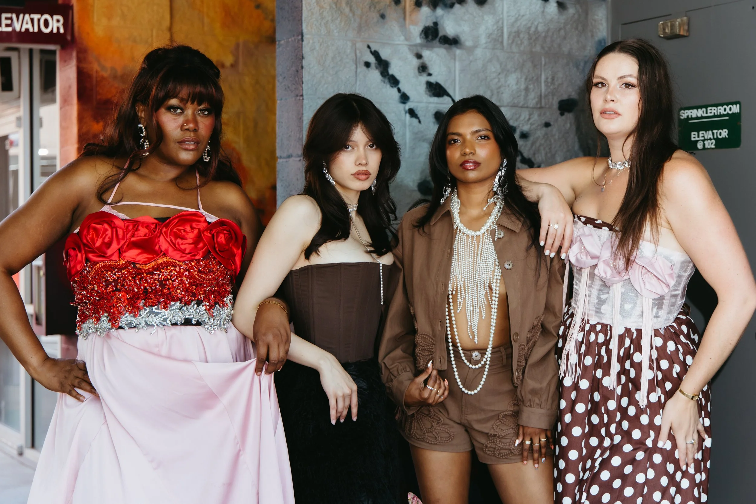 Four women standing together, dressed in colorful and fashionable outfits, posing for a photo indoors near a wall with blue and orange tones, and a sign indicating an elevator and sprinkler room.