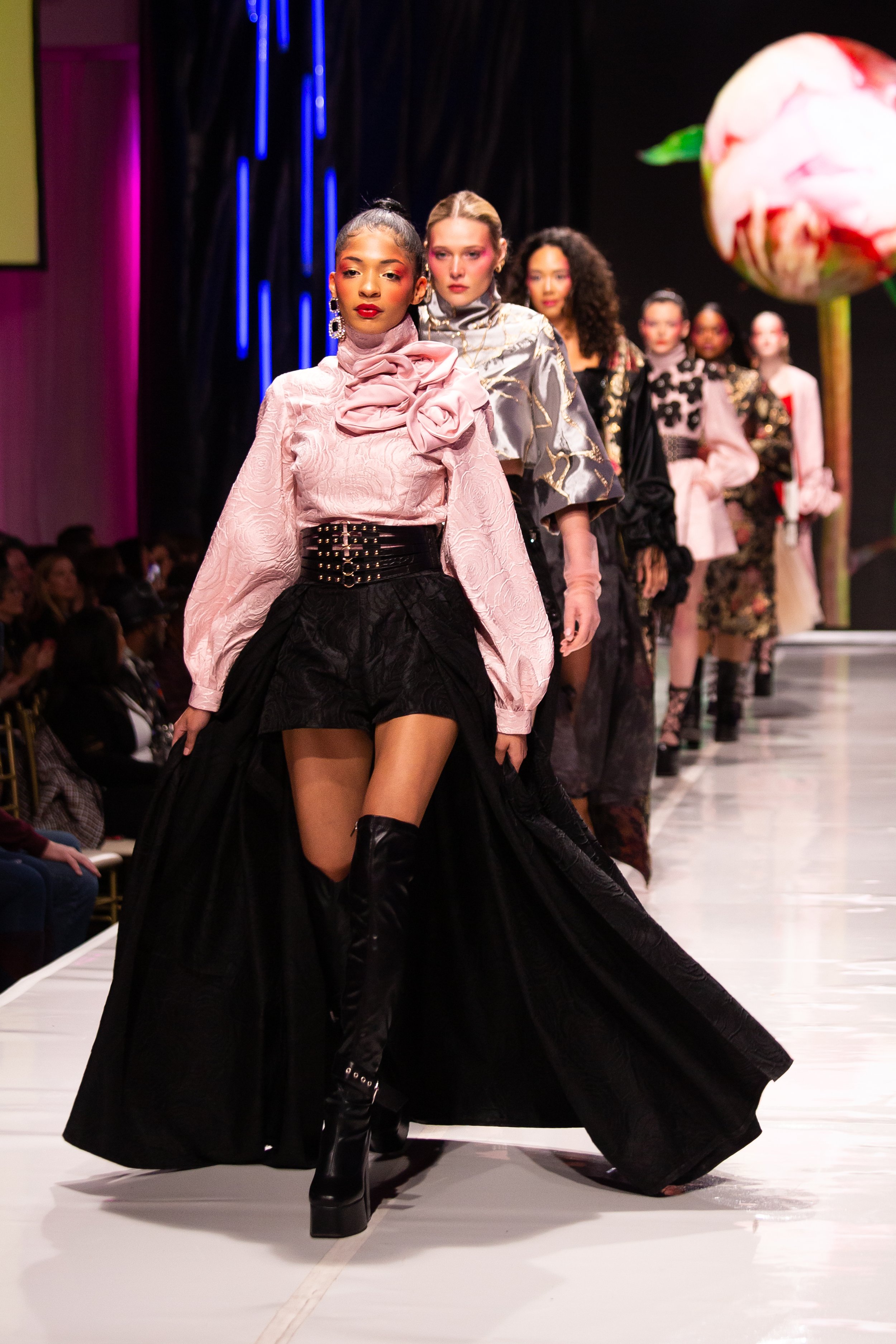 Models walk down runway during a fashion show, wearing high fashion clothing with bold patterns and accessories in a brightly lit venue.