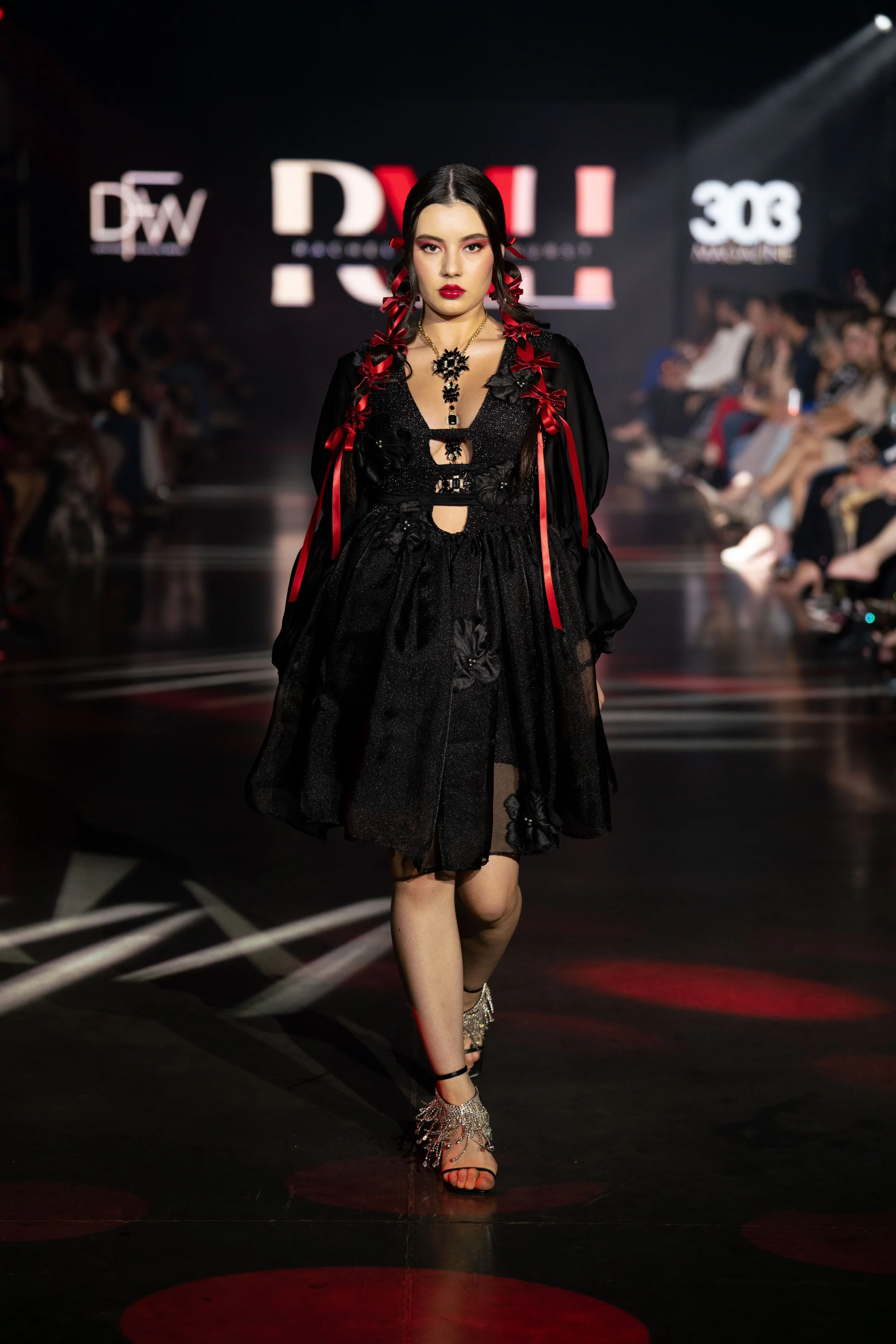 A female model walking down a runway at a fashion show, dressed in a black, sheer, layered dress with floral embellishments, accessorized with jewelry, and wearing strappy heels. The background displays a lit runway with multiple audience members and
