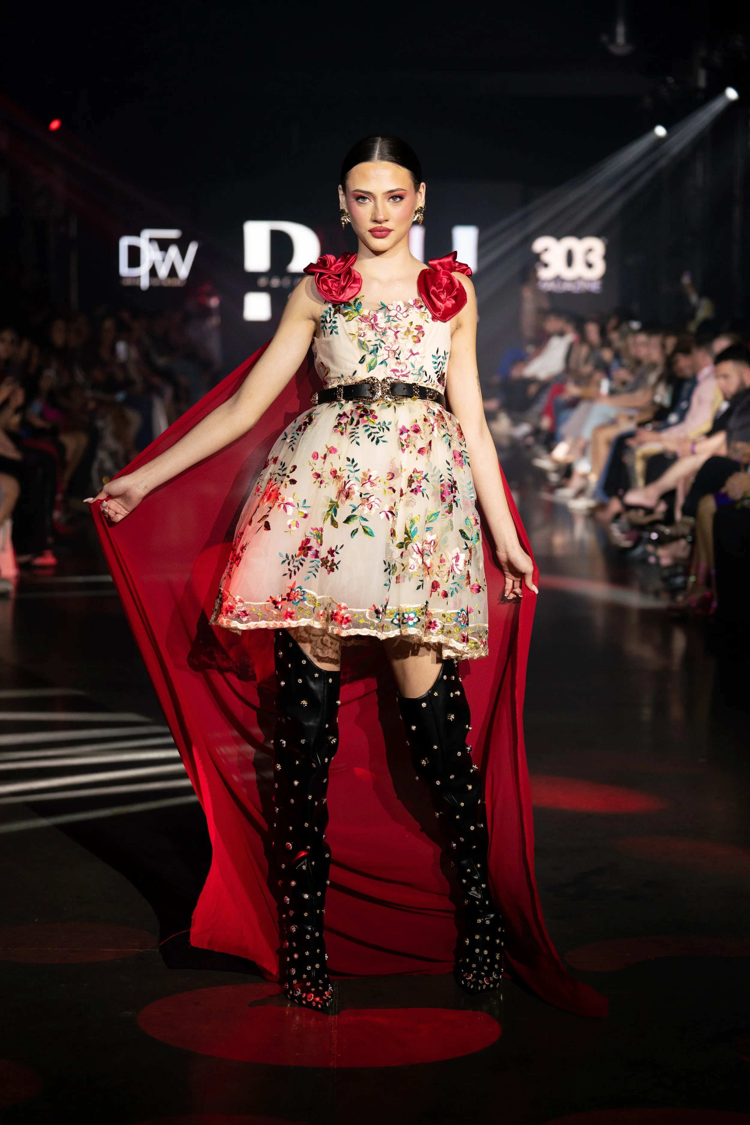 Model walking on runway at fashion show wearing floral dress with large red right shoulder bows, black thigh-high boots with embellishments, and a red cape.