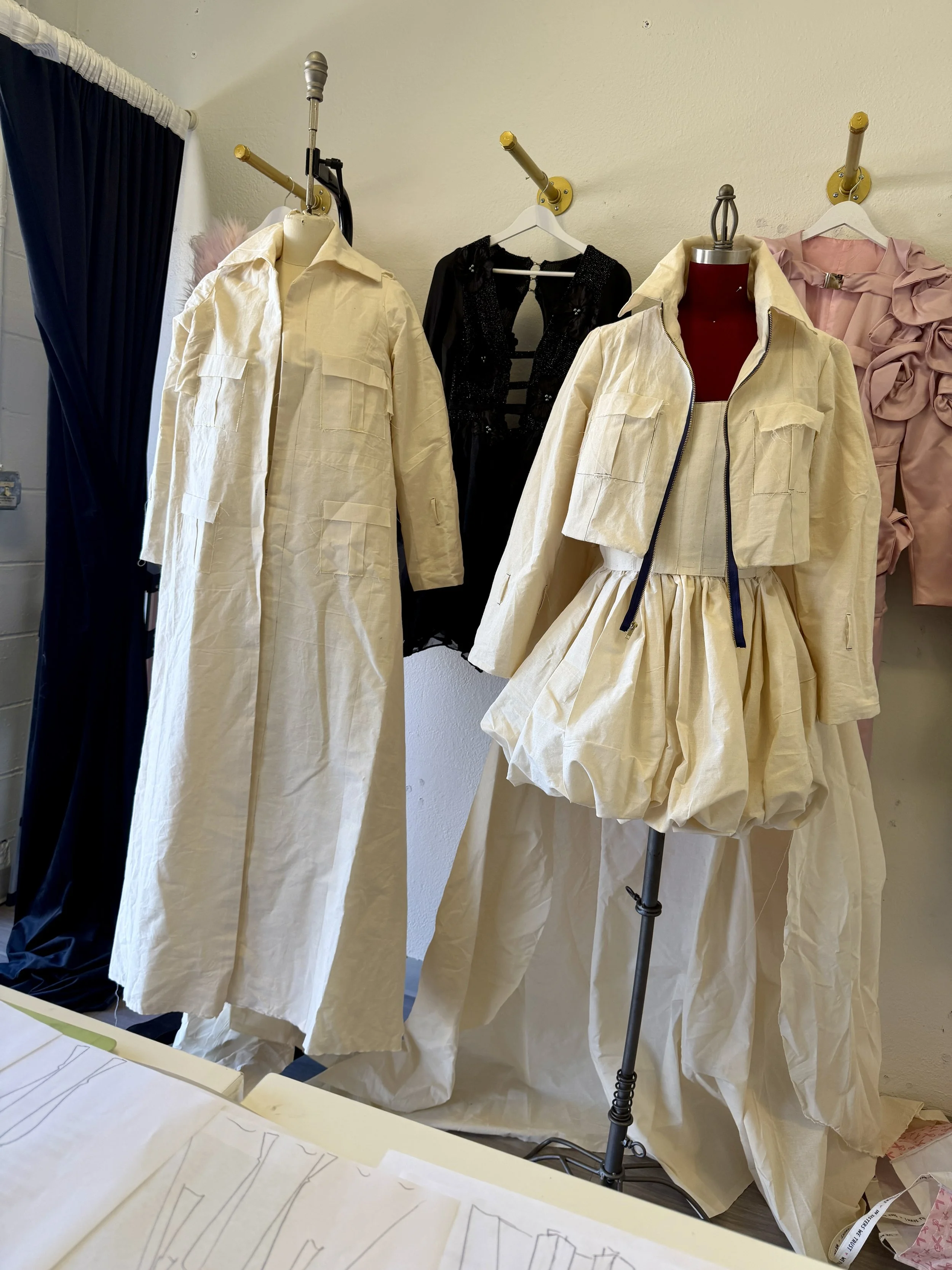 Clothing store display with two cream-colored jackets on mannequins, a black dress in the background, and a pink ruffled garment to the right. There are also sketches or patterns on paper on a table in the foreground.