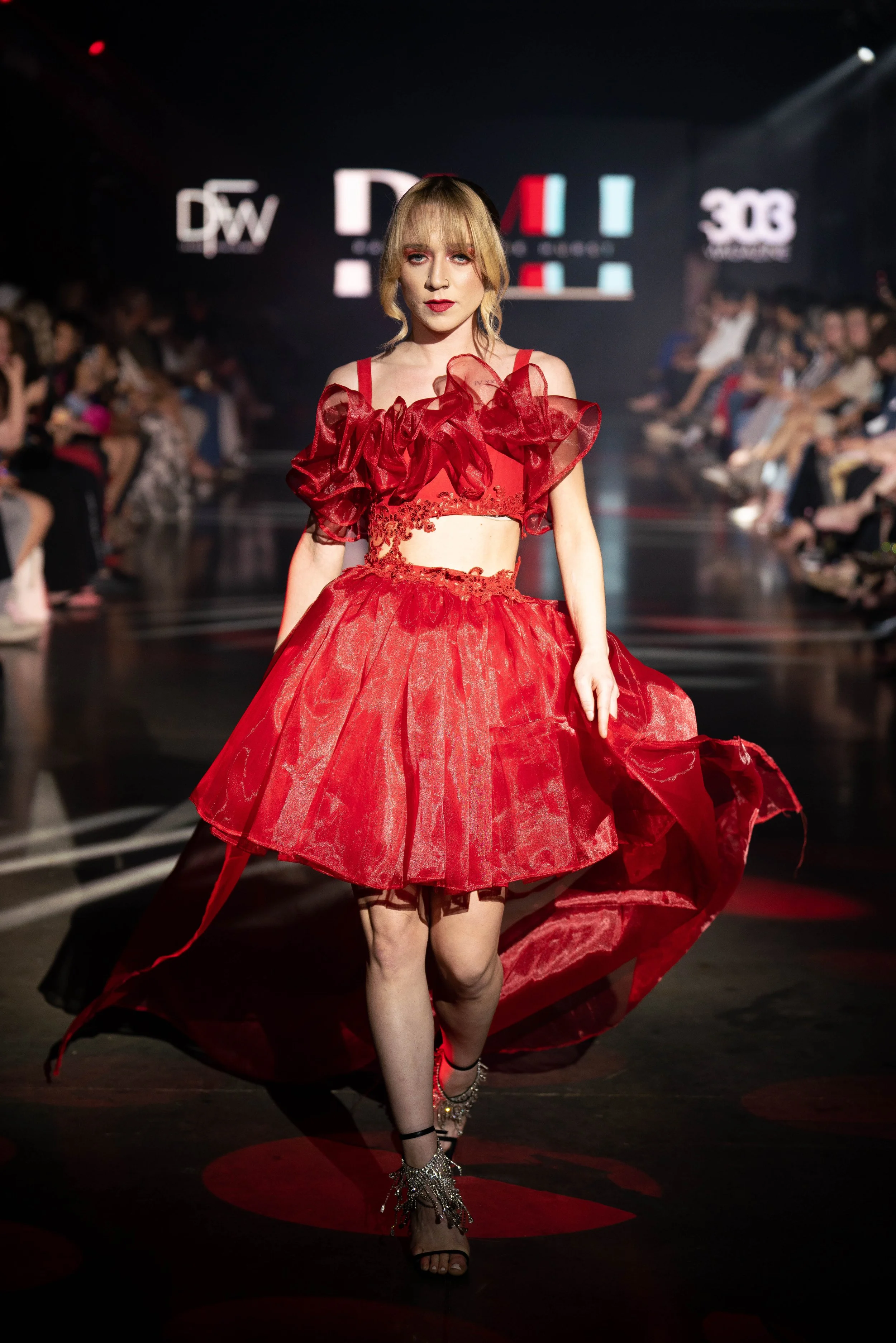 Female model walking on runway in a red dress with ruffled sleeves and sheer skirt at a fashion show.