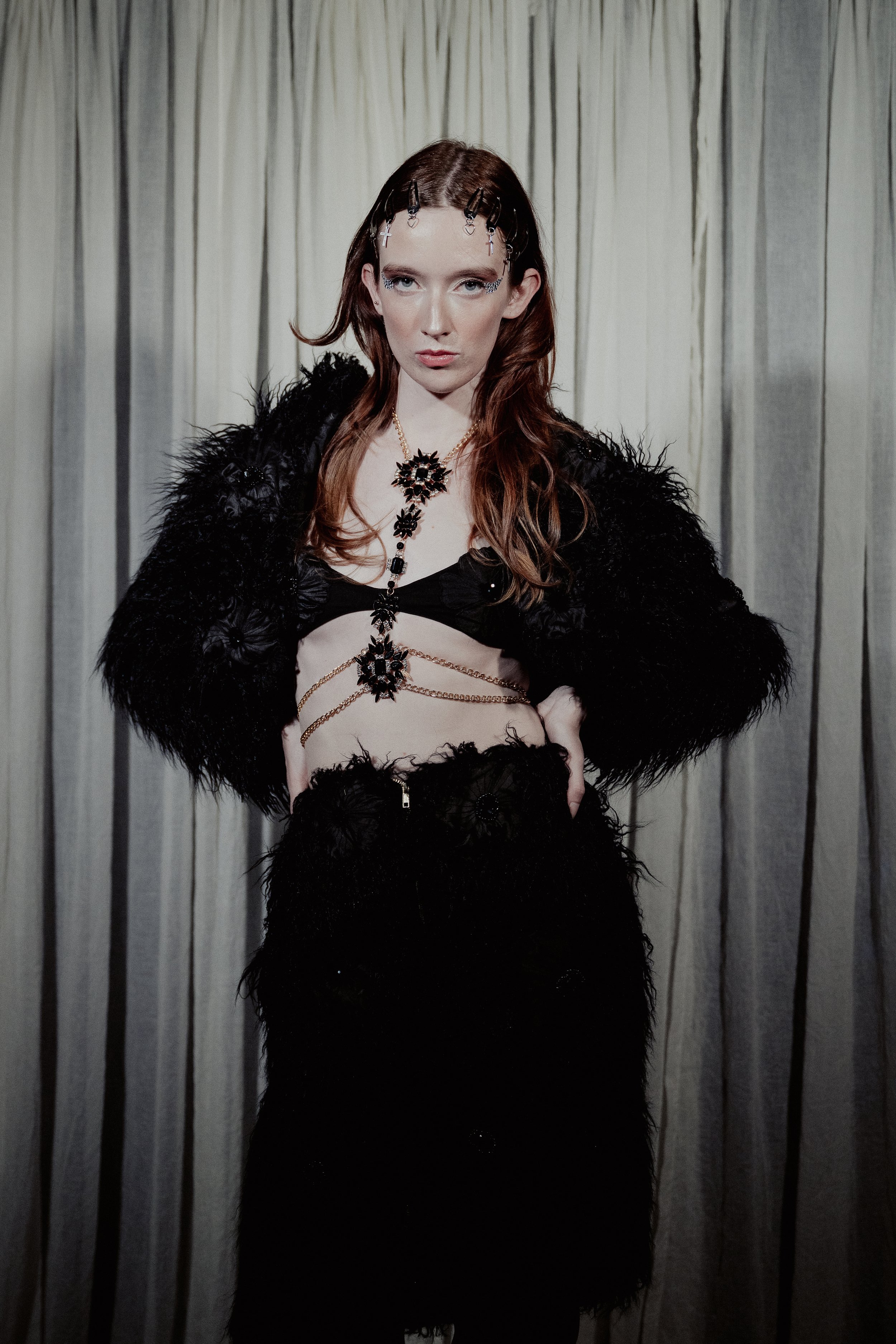 A woman with long red hair and blue eyes, wearing black feathered clothing and jewelry, standing in front of a light-colored curtain.
