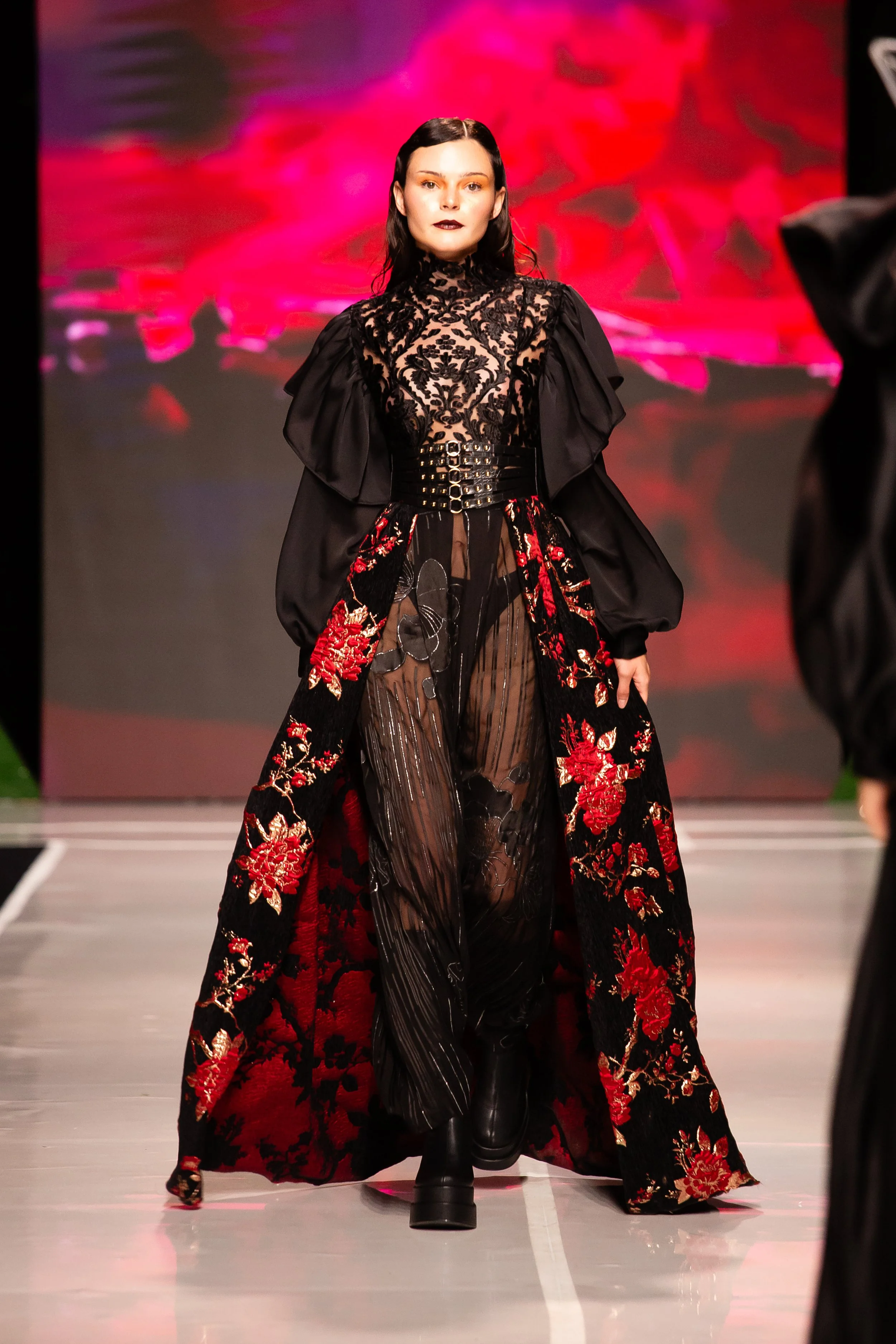 Model walking on runway wearing black and red floral embroidered coat, sheer black pants, and black platform boots with dark makeup and sleek black hair. Red and pink abstract background.