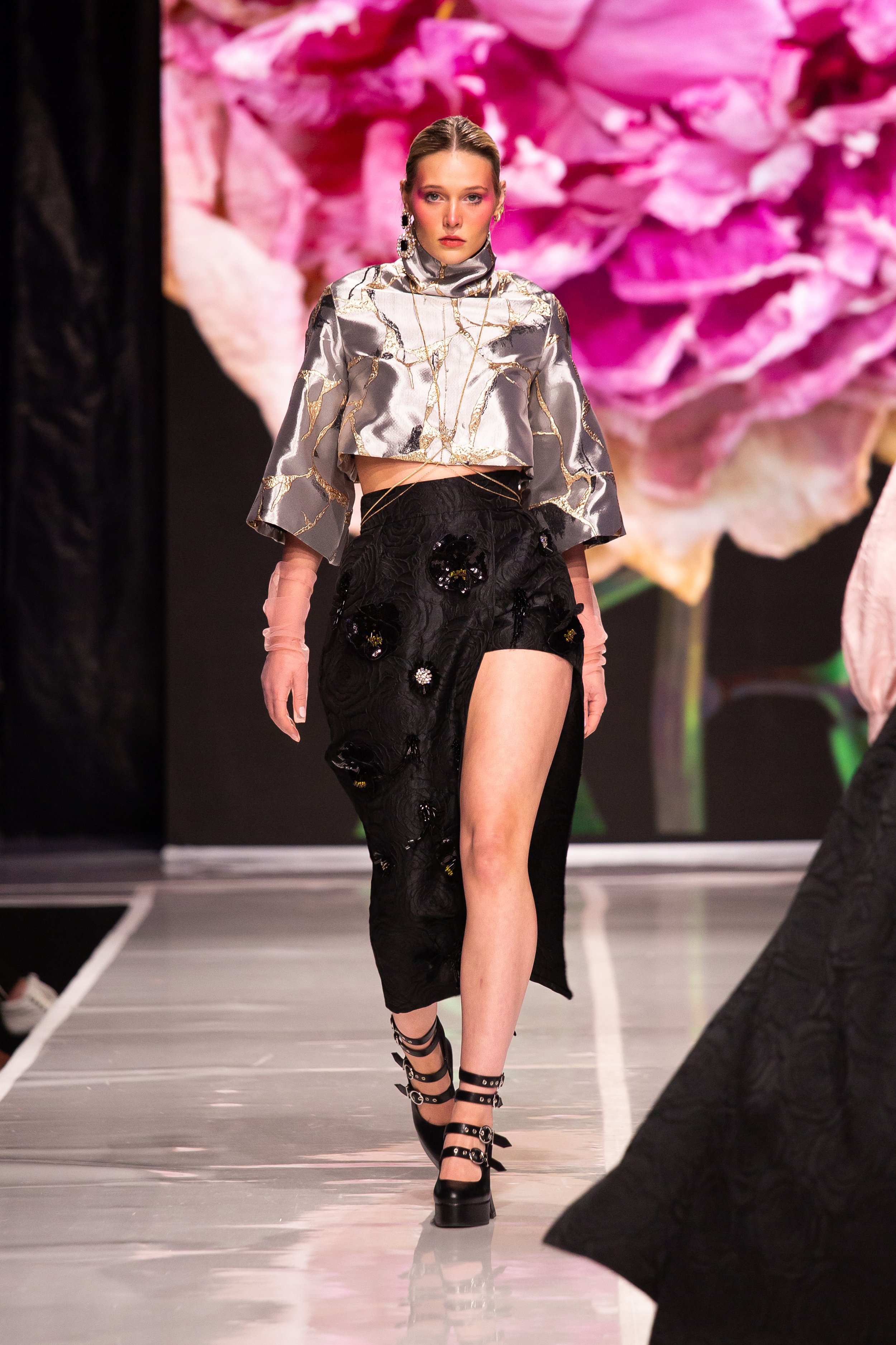 A female model walking on a runway during a fashion show, wearing a metallic cropped top with a gold and black marbled pattern, black textured pants with floral embellishments, and strappy high-heeled shoes. The background features a large pink flowe