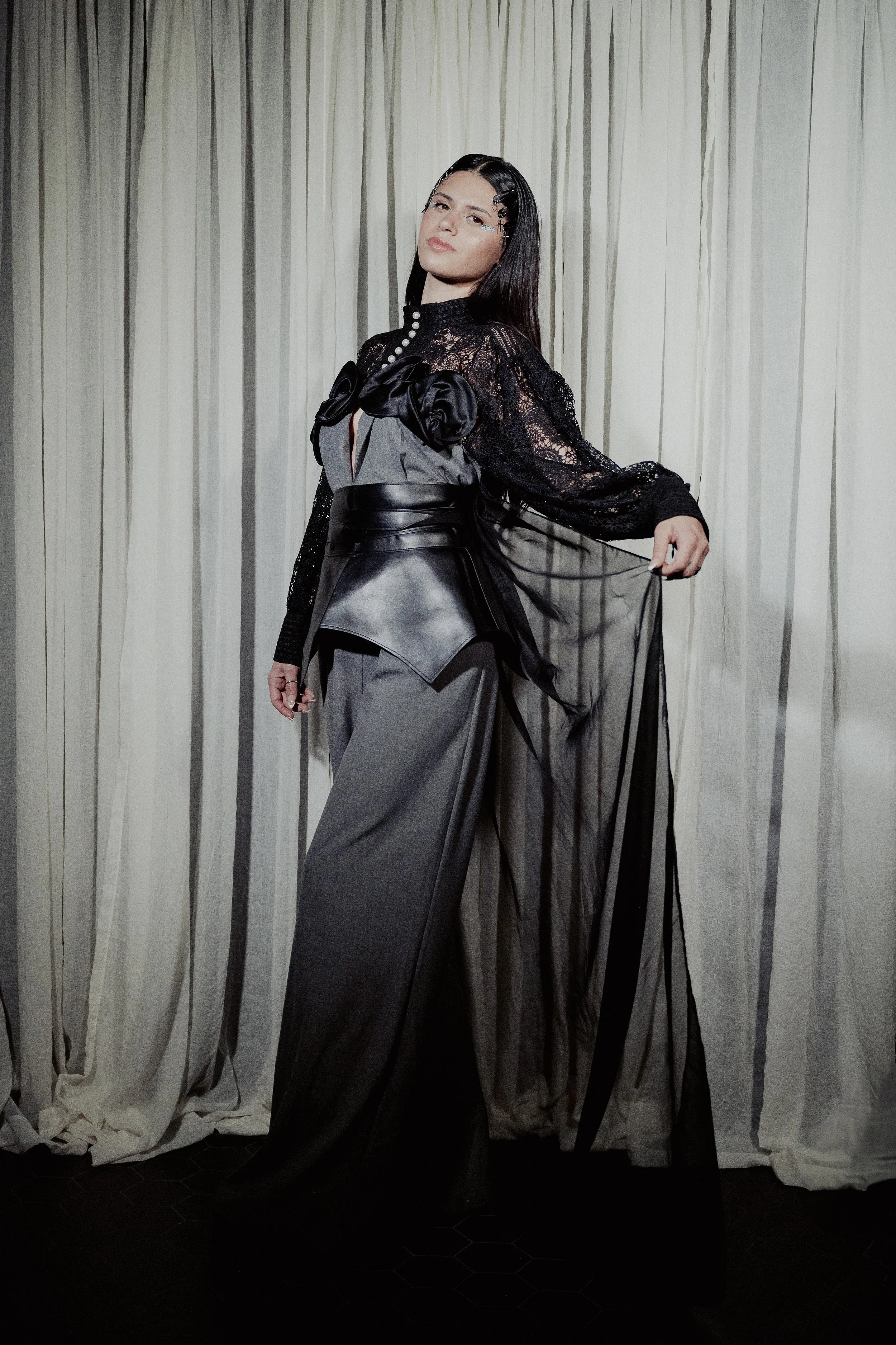 A woman standing in front of a cream-colored curtain, wearing a black lace top, gray wide-leg pants, and a sheer black cape, with dark hair styled straight and a confident expression.