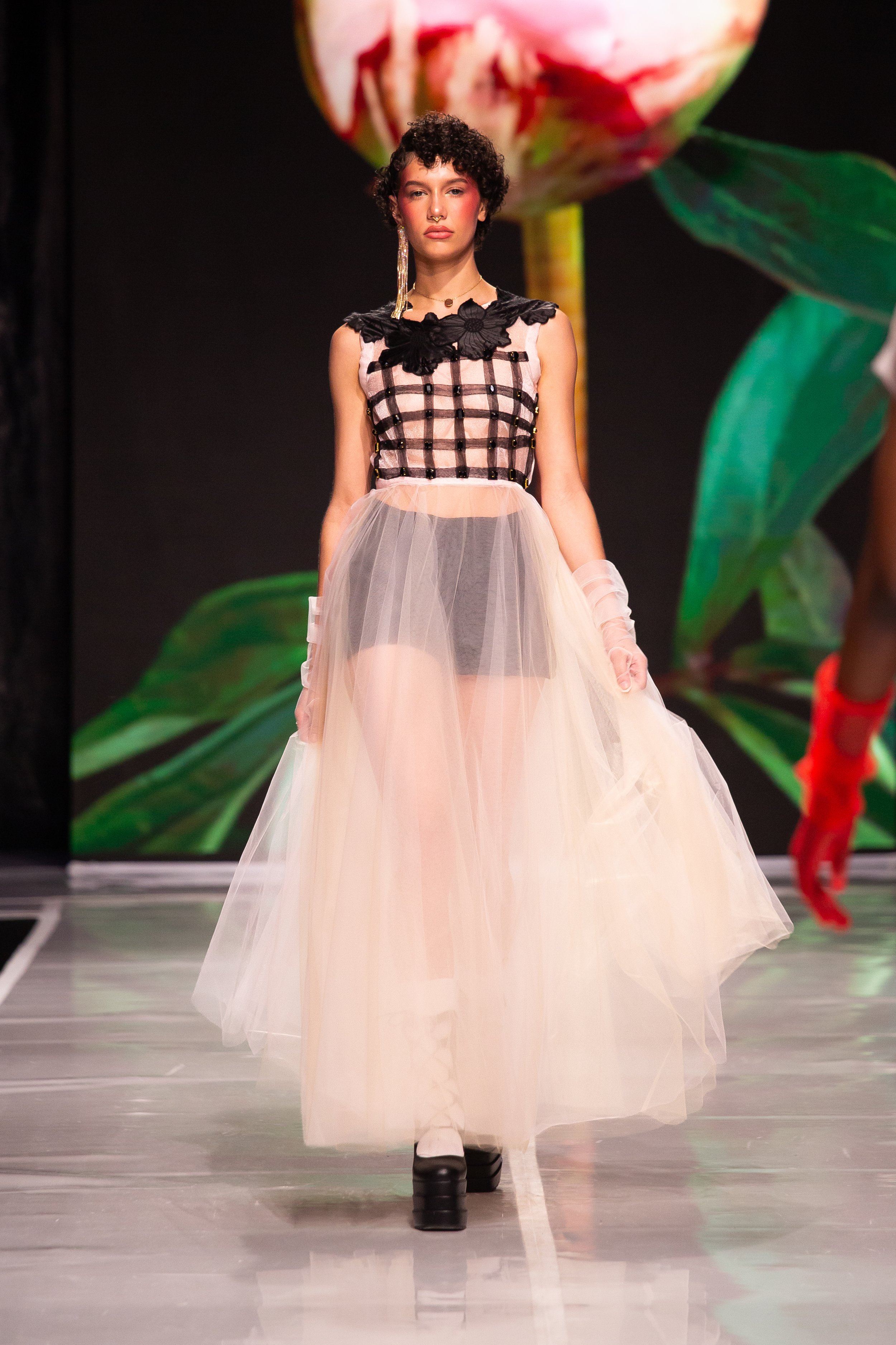 A fashion model walking on a runway, wearing a layered, light-colored tulle skirt and a sleeveless top with black and pink checkered pattern and black floral embellishments. She has short, curly hair, large earrings, and dark shoes. In the background