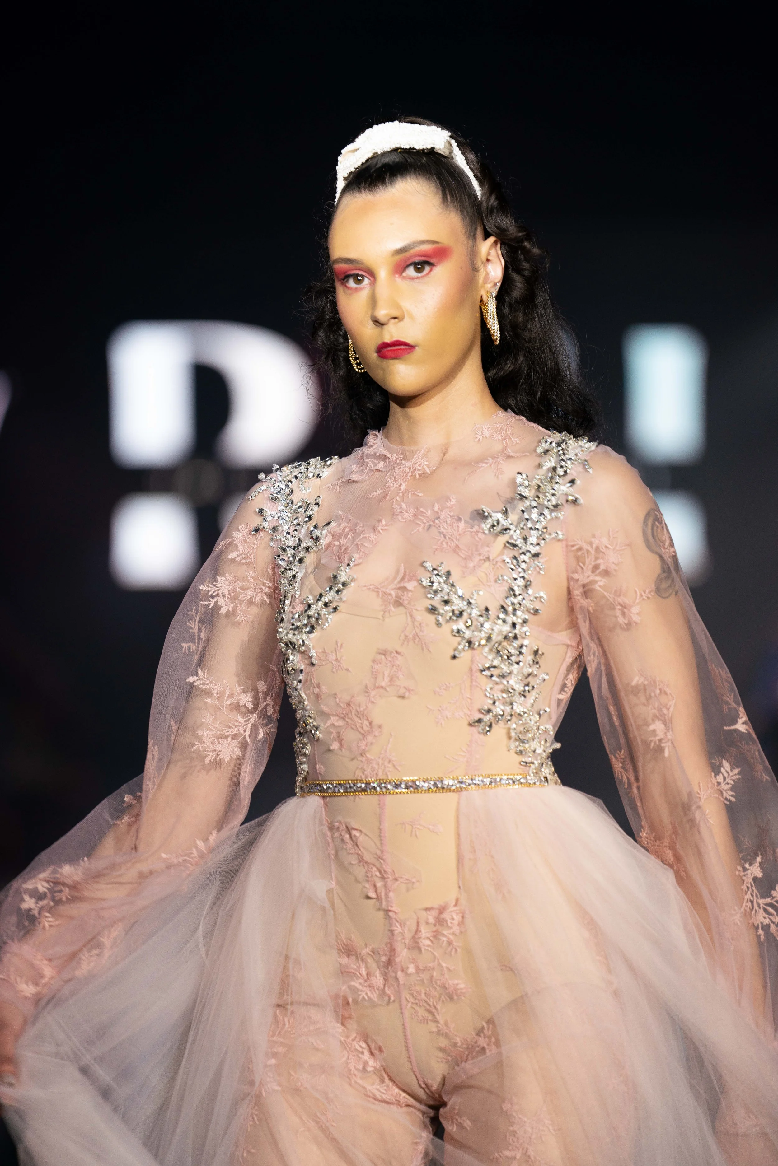 Model with dark wavy hair styled in a high ponytail, wearing a light pink gown with embroidered floral details and sheer tulle layers, accessorized with gold hoop earrings and a white headband, walking on a runway with blurred background lights.