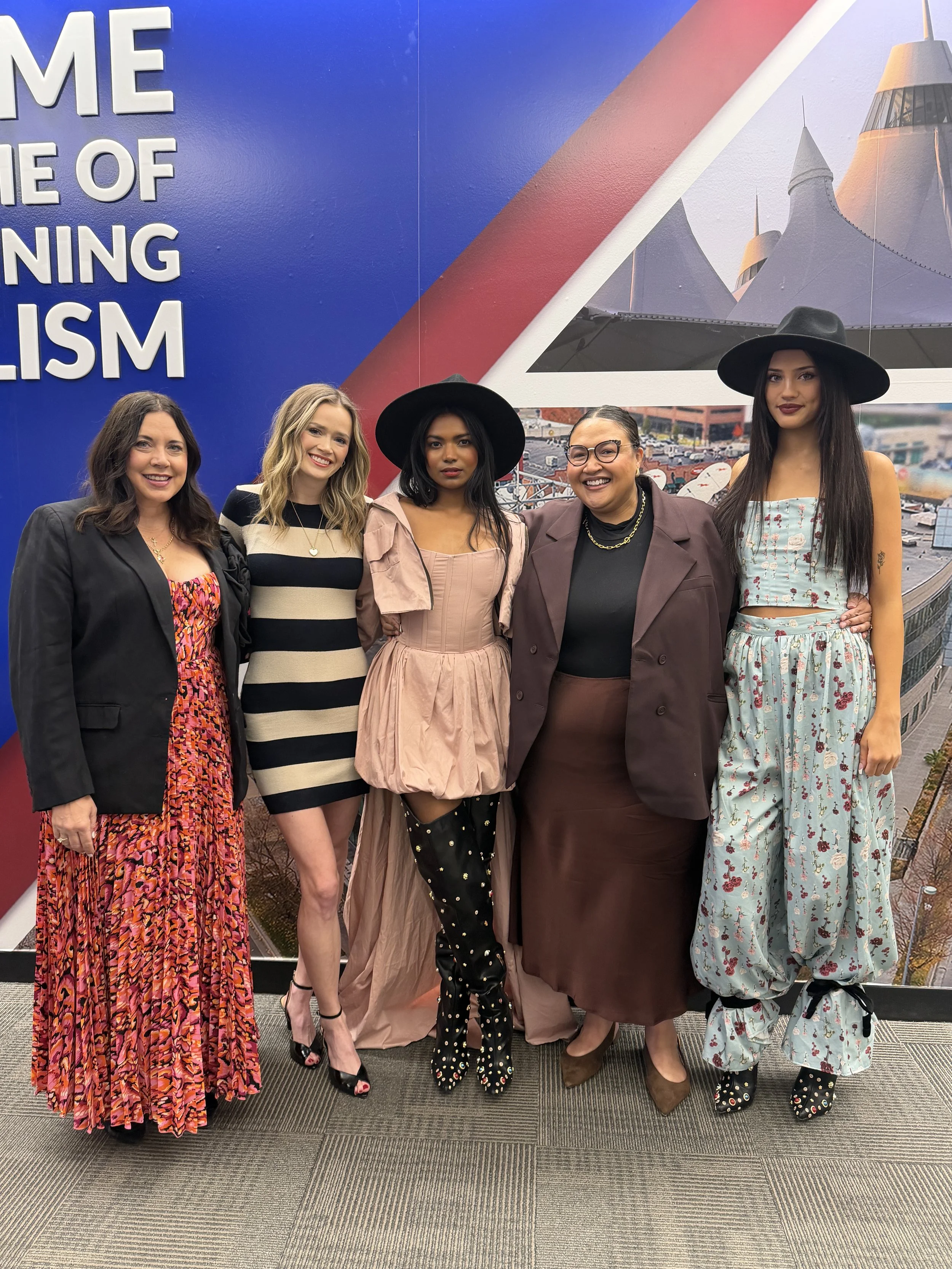 Group of five women standing together in front of a colorful mural with bold text and abstract structures, all dressed in fashionable clothing and accessories.