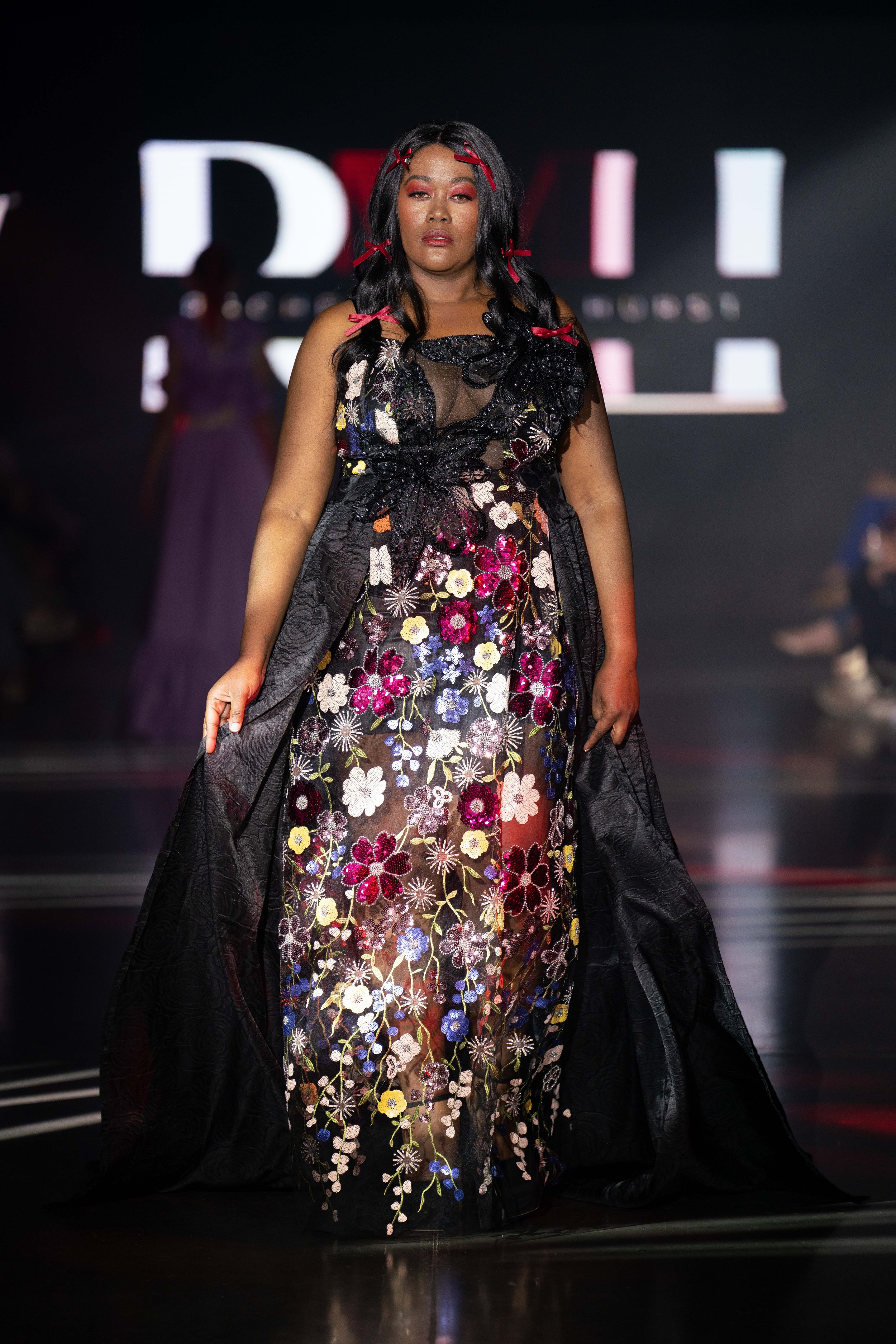 A woman walking on a runway wearing a floral embroidered dress with a sheer black top and black outer layer, with her hair styled in loose waves and adorned with red hair clips. The background displays illuminated letters, and the runway is dark with