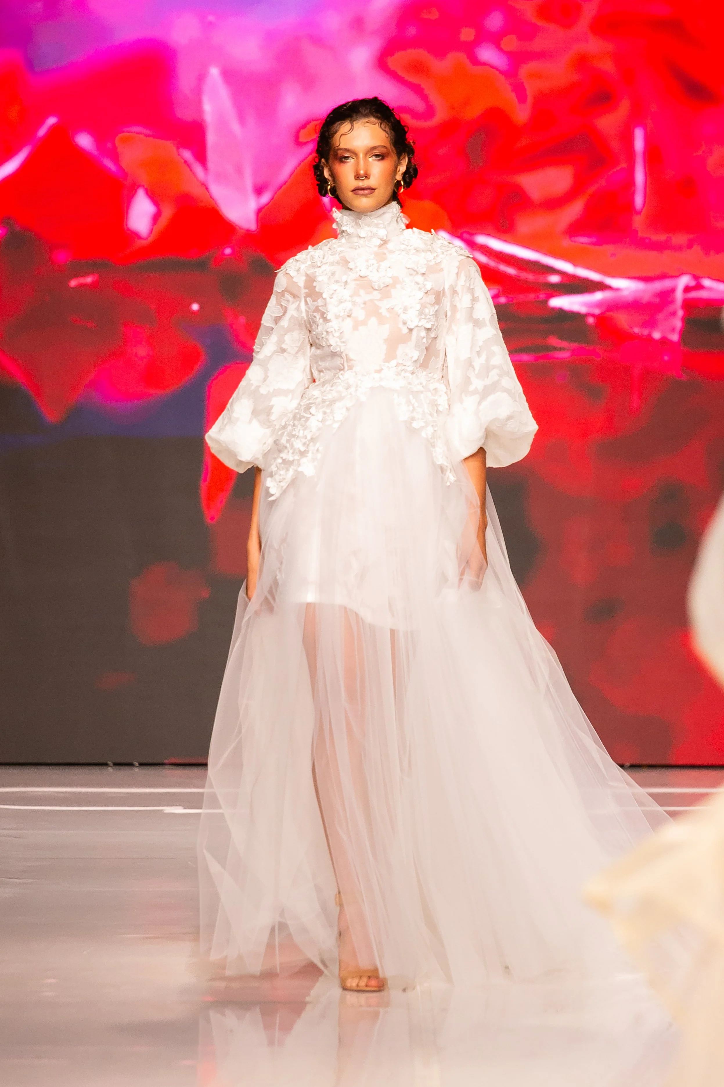 Model walking on runway wearing a strapless white bridal gown with floral lace appliqué, sheer tulle skirt, and high heel sandals, against a vibrant red and pink abstract background.