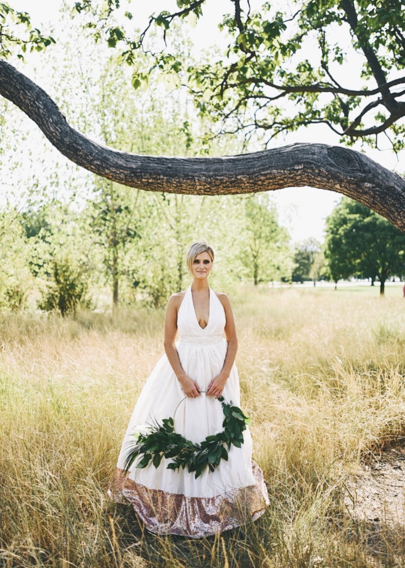A woman in a white dress standing in a sunny, grassy field holding a leafy wreath, with a large tree branch overhead and other trees in the background.