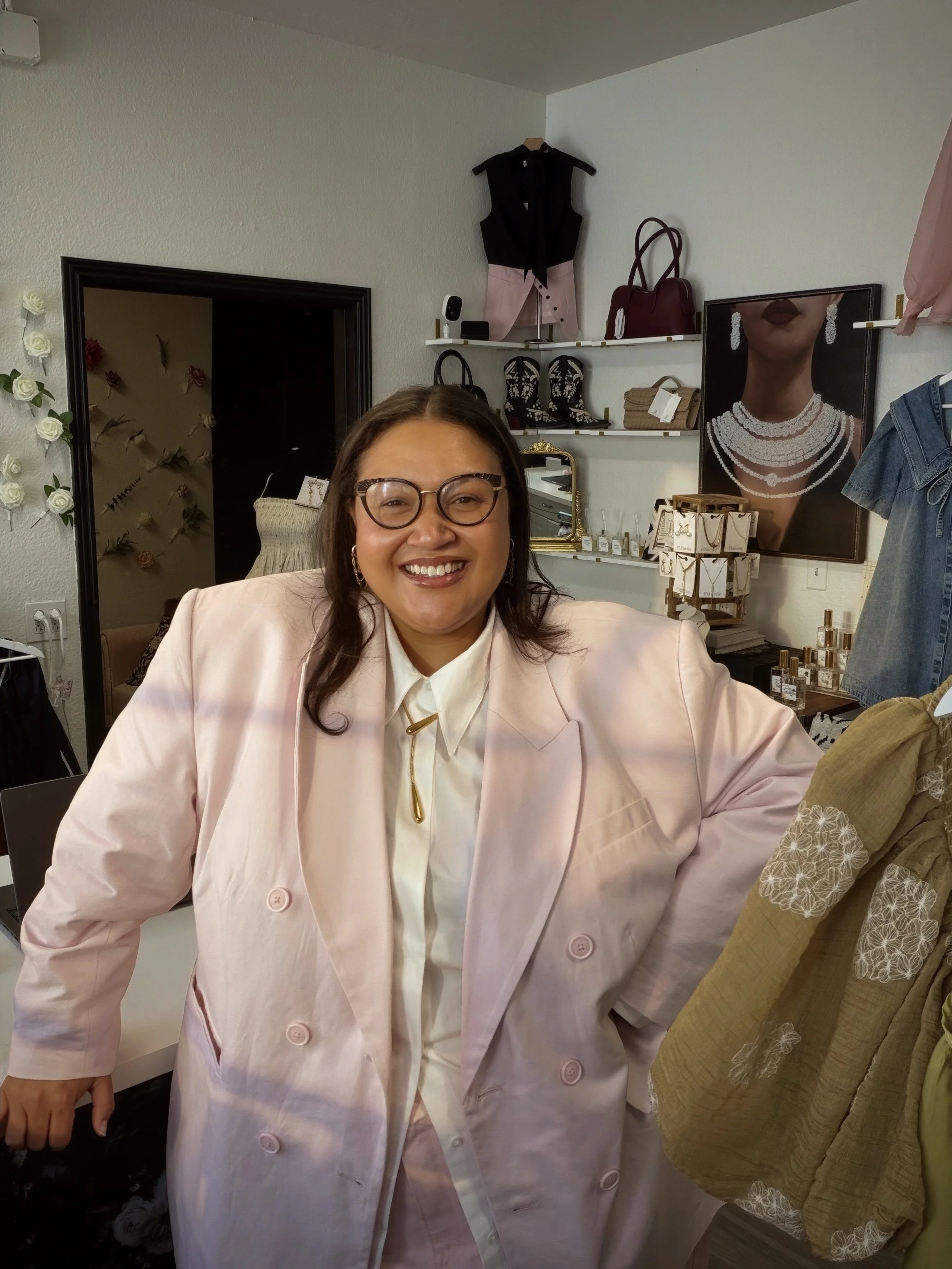 A woman smiling and wearing glasses, dressed in a light pink blazer over a white shirt, standing in a store with shelves displaying handbags, shoes, and jewelry, and a framed portrait of a woman wearing pearl jewelry behind her.