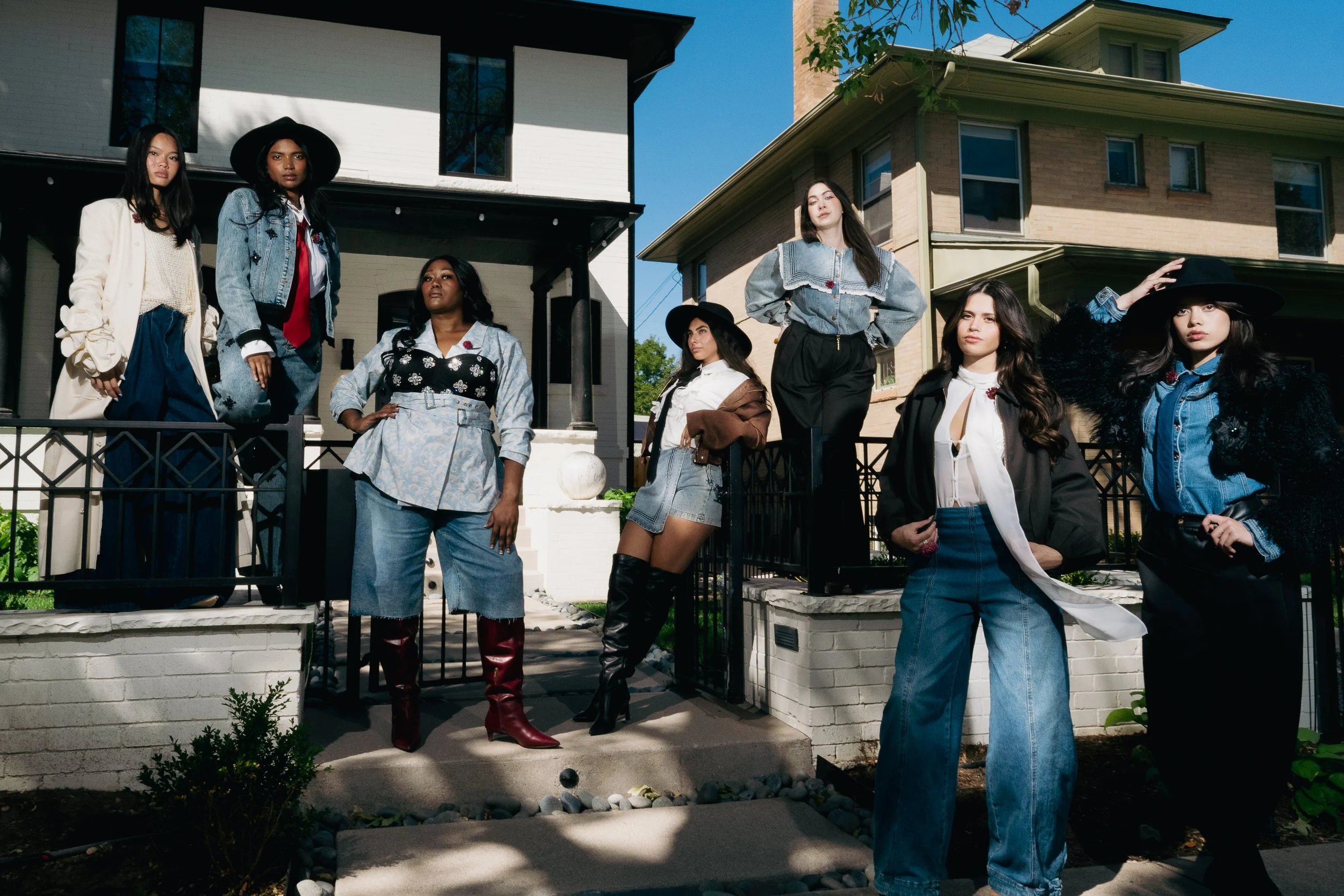 Group of seven women standing outside in front of two houses, dressed in stylish outfits, with serious expressions.
