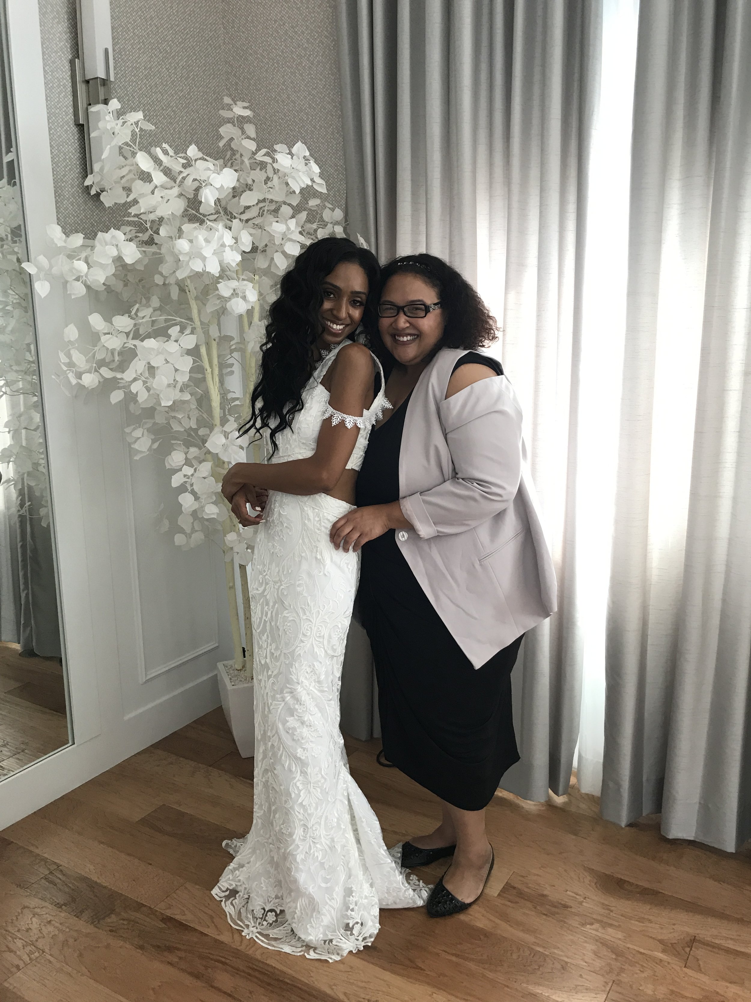 Two women standing close together and smiling, one wearing a white wedding dress and the other wearing a light pink blazer over a black dress, in front of a window with curtains.
